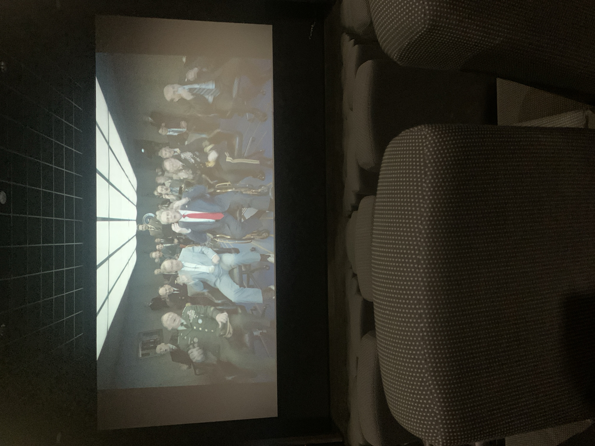 In a dimly lit, modern movie theater, the large screen displays a scene of men in formal attire, some in military uniforms, seated and appearing engaged. The foreground is dominated by rows of dark, upholstered theater seats, some appearing to be recliners. The environment suggests a viewing of a film or presentation. There is no indication of the location being in Sinalunga, Italy.