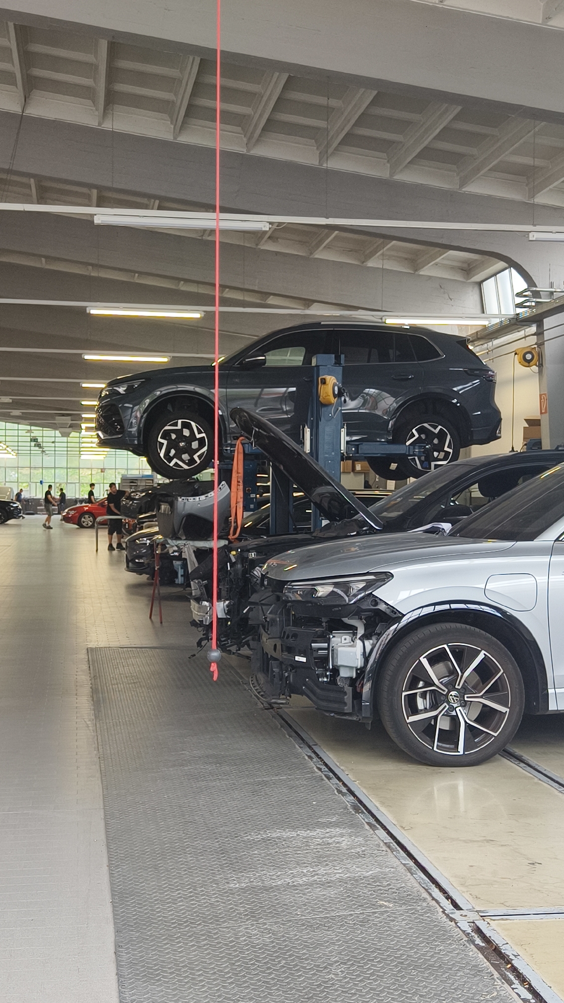 This is a vertical, eye-level, medium shot of a car repair shop in Vienna, Austria. In the foreground, the front of a silver car is visible, with its bumper removed. To the left, a dark-colored car with its hood open is parked on a ramp. Further back, a dark gray SUV is elevated on a car lift. Several other cars are parked in the background, and some people are standing around them. The environment appears to be a well-lit indoor space with a high ceiling and industrial lighting. The floor is a combination of grey tiled and metal diamond plate surfaces. A red laser line is visible hanging from the ceiling, likely used for alignment purposes.