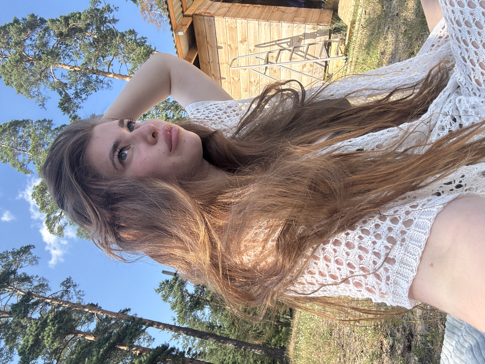 In this media file, a young woman with long, reddish-brown hair is captured in a close-up shot, with her face turned towards the camera. She is lying down outdoors, with her arm bent and hand behind her head, suggesting a relaxed or pensive pose. Her expression is soft, with her eyes looking slightly upwards and to the side. She is wearing a white, intricately patterned knitted garment, possibly a top or a dress, with a lace-like design.
The background reveals an outdoor setting with elements that provide context. Tall pine trees with green needles dominate the upper left portion of the frame, with a clear blue sky visible through their branches, indicating daylight. To the right, a wooden structure, possibly a small cabin or shed, is visible, constructed from light-colored wooden planks. A metal railing or ladder is attached to its side. The ground is covered with dry grass and some scattered white flowers, suggesting a natural, possibly rural or park-like environment. The overall lighting and the sun's position, casting shadows from the wooden structure, indicate that the photograph was taken during the daytime, likely in the late morning or afternoon. The weather appears to be sunny and pleasant.
The image is oriented in a vertical, sideways perspective, requiring the viewer to turn the image to see it correctly. No visible text is present in the media file. The overall mood conveyed is one of tranquility and natural beauty, with the woman appearing at ease in her surroundings.