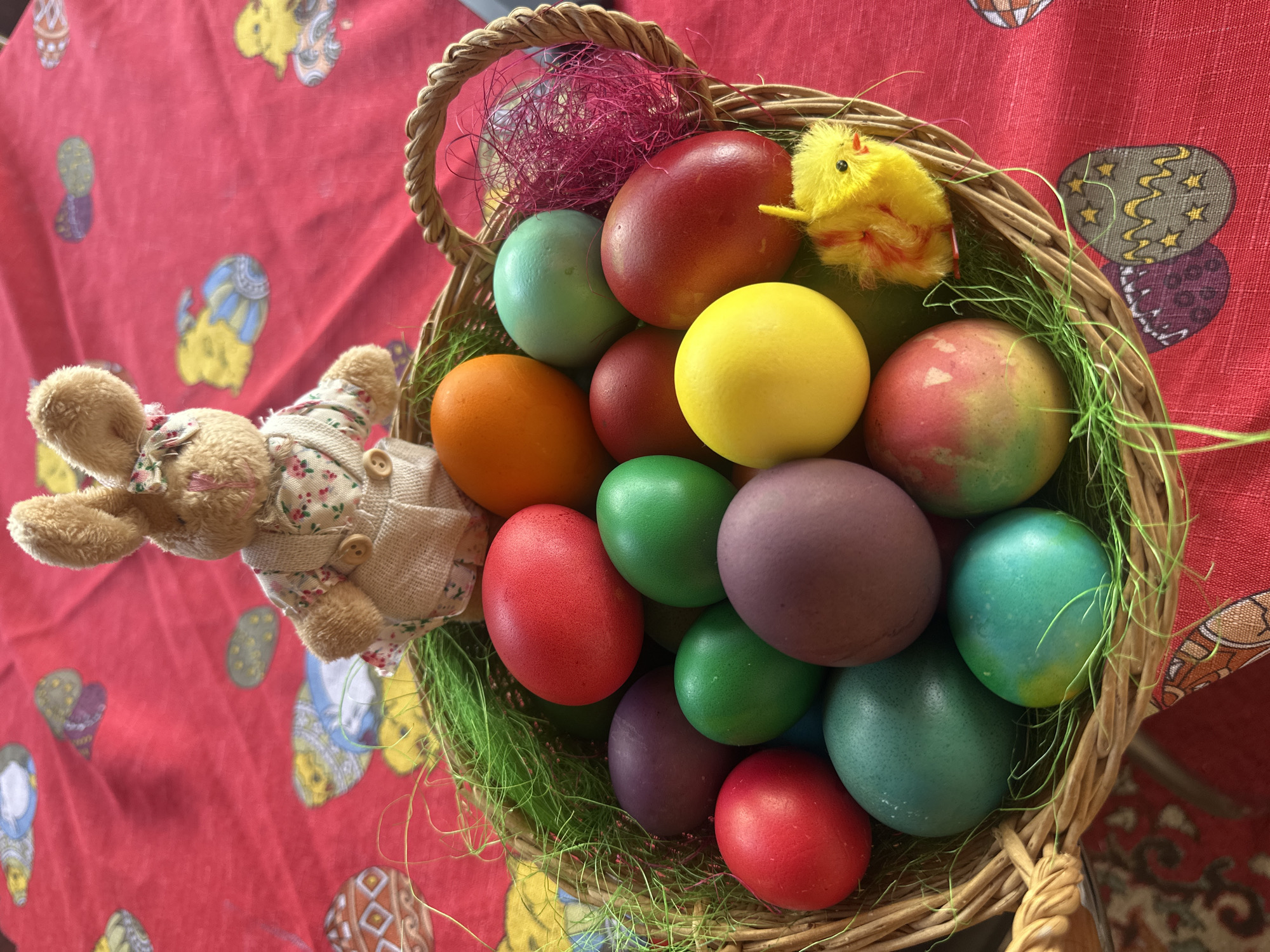 A high-angle shot captures a woven basket overflowing with vibrantly colored Easter eggs. The eggs, a variety of hues including red, orange, yellow, green, purple, and teal, nestle within a bed of artificial green grass and a touch of pink synthetic material. A stuffed bunny, dressed in a floral-patterned shirt and beige jacket, appears to lean against the basket's edge. Adjacent to the eggs, a small, fuzzy yellow chick adds to the Easter theme. The basket rests on a red tablecloth adorned with various Easter-themed patterns. The location of this scene is Unknown, Unknown.