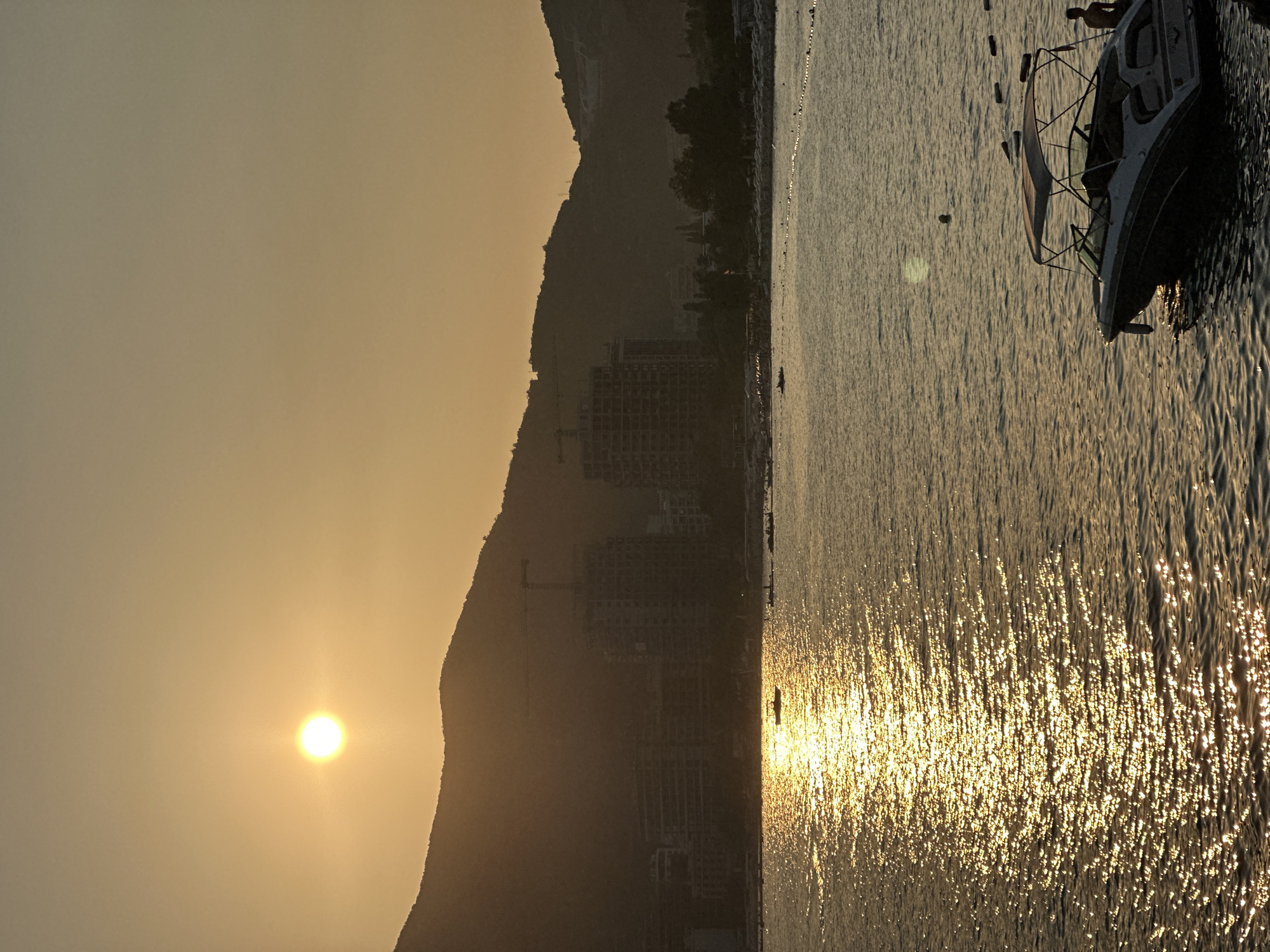 This is a vertical, medium shot photograph of a coastal scene at sunset, likely in Budva, Montenegro. The sun, a bright, hazy disc, is low in the sky, casting a warm, golden glow across the water and silhouetting the mountainous terrain behind. The water is rippling, with the sun's reflection creating a shimmering, broken path of light.
In the middle ground, a crescent-shaped beach is visible, lined with beach chairs and umbrellas, though many are empty. Behind the beach, a line of buildings, some appearing to be under construction with cranes visible, forms the urban backdrop. Lush green trees are scattered amongst the buildings.
In the foreground, a motorboat with a canopy is moored to the right, its hull dark against the illuminated water. A person stands near the stern of the boat. Further out on the water, a small boat with two occupants is visible, silhouetted against the bright reflection of the sun. Another person is swimming in the water to the left of the motorboat. The overall mood is tranquil, with the warm light suggesting the end of a summer day.