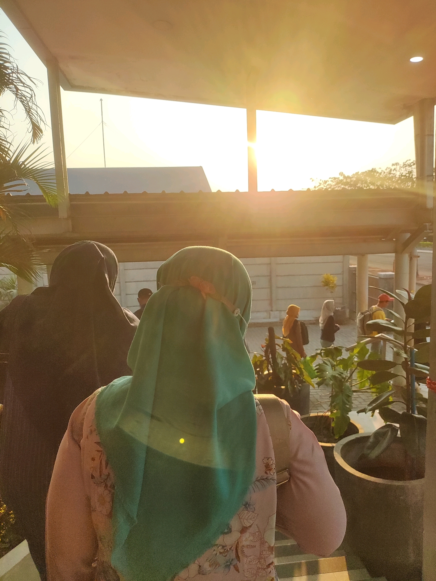 In Telagamurni, Indonesia, at golden hour, a group of individuals are seen walking outdoors near a building with a wide, covered overhang. The sun is low in the sky, casting a warm glow and lens flare across the scene. In the foreground, two women are facing away from the camera, one in a dark hijab and the other in a teal hijab with a pink accent. Behind them, in the middle ground, several other people are visible, including a man wearing a red cap and a yellow shirt, and two women in hijabs. The environment includes potted plants and a concrete structure. The overall atmosphere is peaceful, with the natural light creating a serene ambiance.