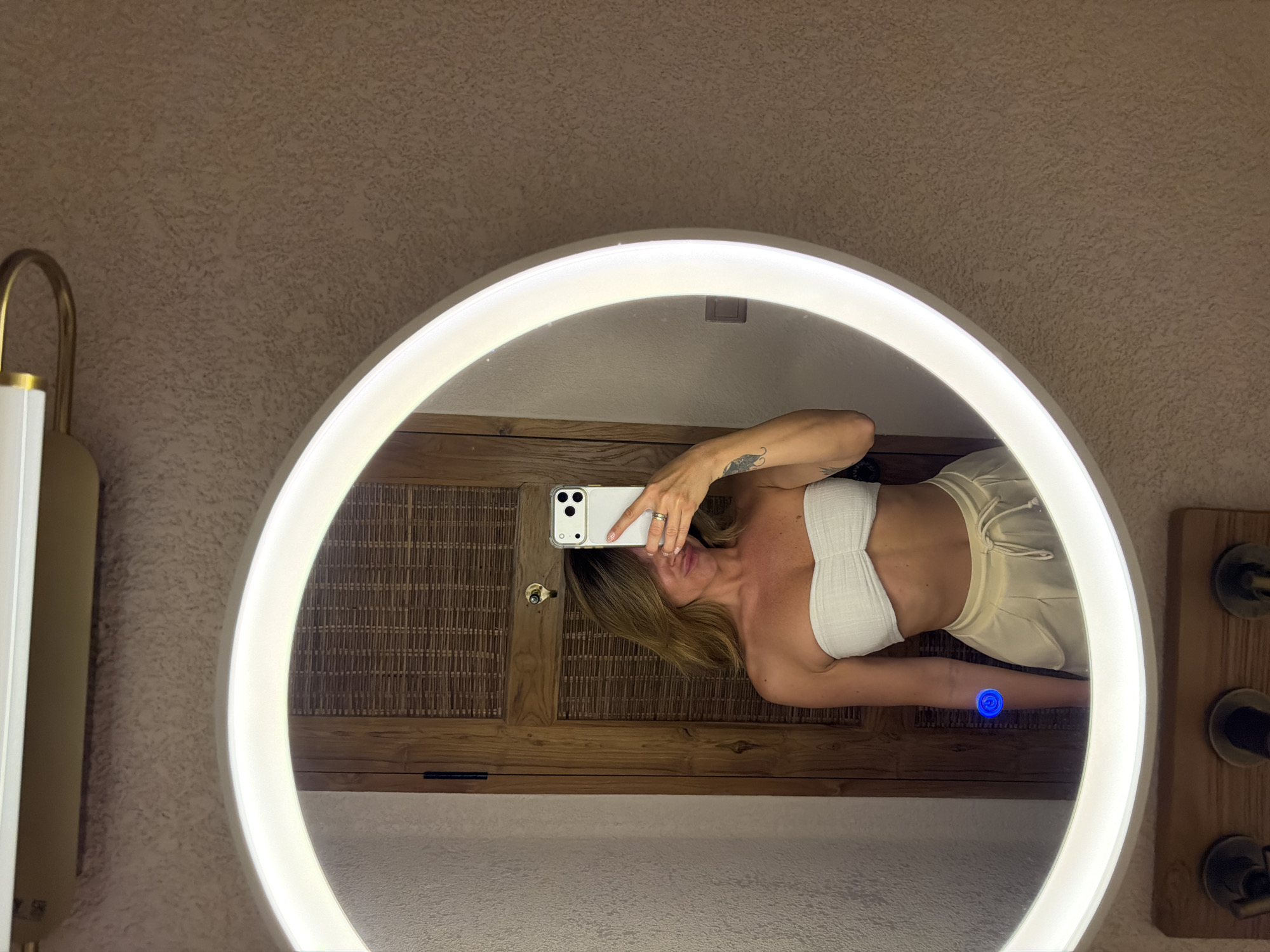 The image displays a woman taking a mirror selfie in a well-lit indoor setting, rotated 90 degrees counter-clockwise. The main subject is the reflection of a woman with long blonde hair, captured horizontally within a large, round mirror framed by a bright, continuous LED light strip.
She is wearing a white strapless top and light-colored shorts or pants, holding a white smartphone with multiple cameras in her left hand (appearing as her right in the reflection) to photograph herself. A ring is visible on her left hand, and a tattoo is discernible on her right upper arm (left arm in the reflection). A prominent blue circular glowing button or indicator is visible on the mirror's surface near her reflected right forearm.
Outside the mirror on the left, a brass-toned light fixture with a white shade is partially visible against a textured, light brown wall. Within the mirror's reflection, behind the woman, dark wooden panels with a woven pattern suggest either cabinetry or a decorative wall. To the right of the mirror, outside its frame, more dark wooden paneling with two faucet handles or knobs implies a vanity or sink area.
The scene suggests a modern bathroom or dressing room, likely in a hotel or resort in Mengwi, Indonesia, given the style and materials. The lighting is artificial, indicating an indoor environment, possibly at night or in a windowless room. There is no visible text in the image.