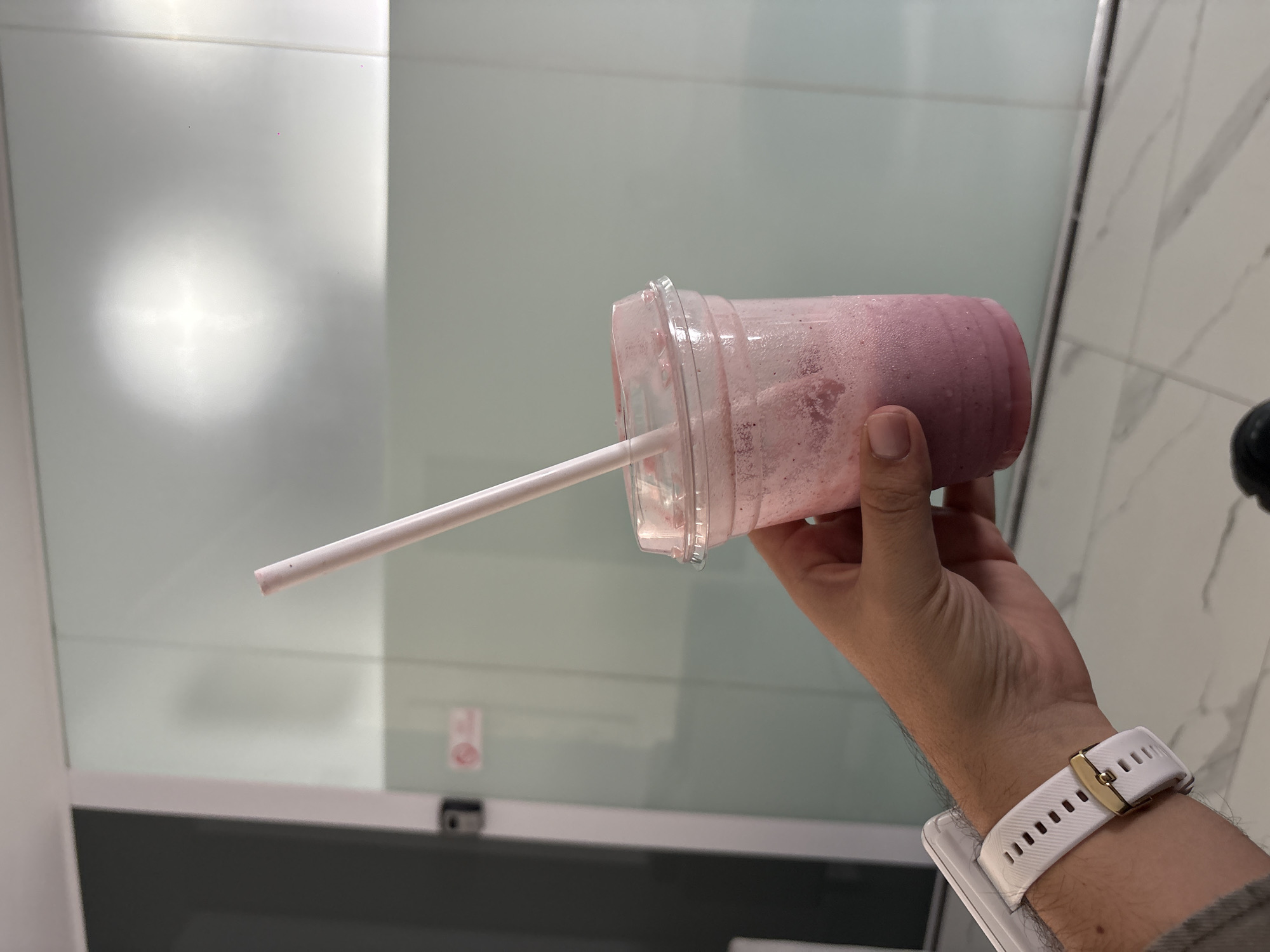 A person holds a partially consumed pink smoothie in a clear plastic cup with a white straw. The person is wearing a white smartwatch on their left wrist. The background consists of a white wall, with the left side of the wall appearing smooth, and the right side resembling marble or tile. The location is not identifiable as Puebla, Mexico based on the provided image details.