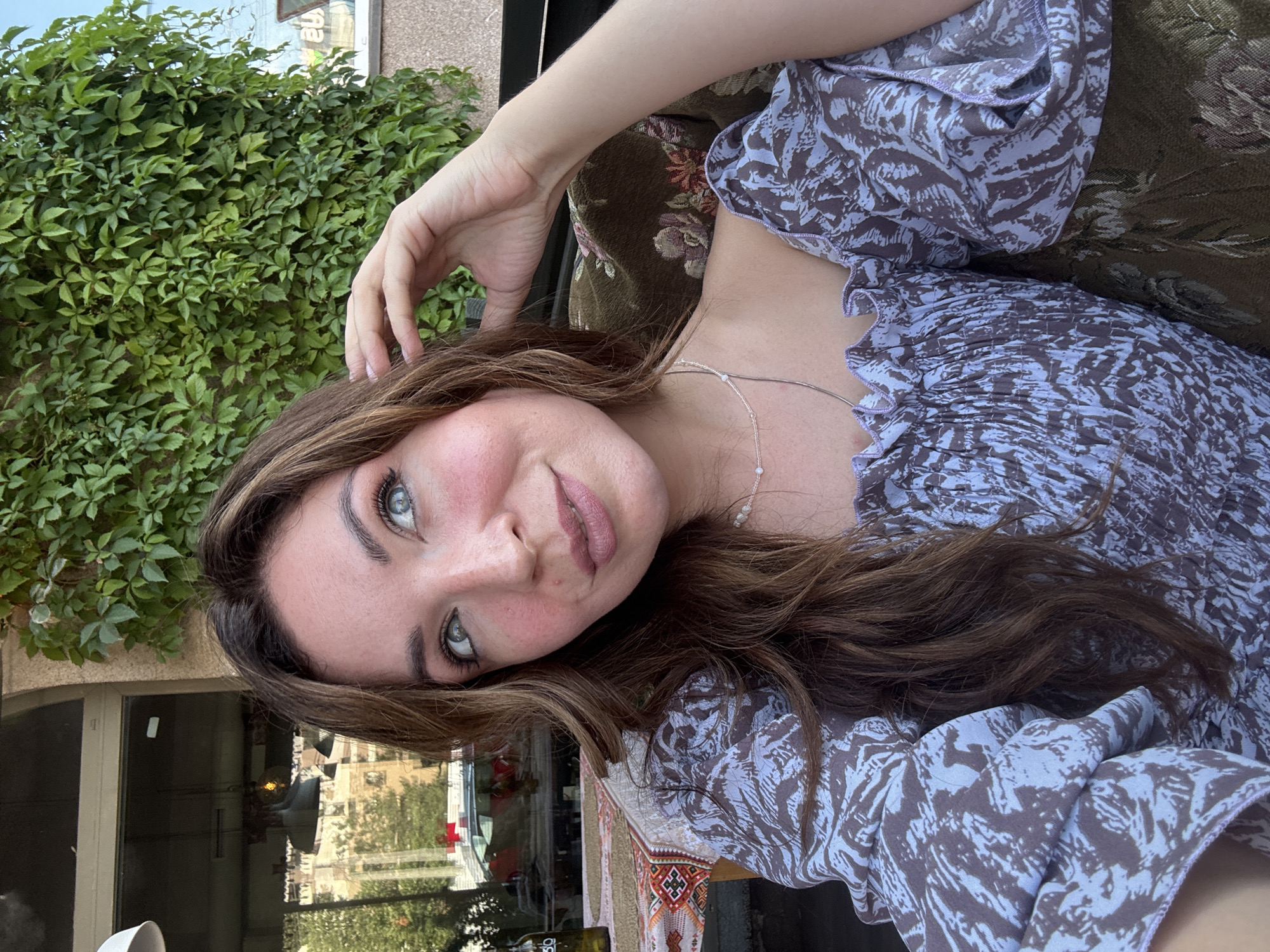 A woman is captured in a close-up shot, seated outdoors. She has long, wavy brown hair, fair skin with blush on her cheeks, and bright blue eyes. Her head is tilted slightly upwards and to the left, with her right hand touching her hair near her temple. She is wearing a light purple off-the-shoulder top with a dark pattern. A delicate pearl necklace is visible around her neck.
The background suggests an urban setting in Kryvyi Rih, Ukraine. To the left, a window with reflections of buildings and a vehicle indicates an interior space, possibly a cafe or restaurant. A bottle is visible on a table covered with a patterned cloth. To the right, a dense wall of green ivy covers a building, suggesting an outdoor patio or courtyard. The lighting is natural, indicating it is daytime. The woman appears relaxed and is looking away from the camera.