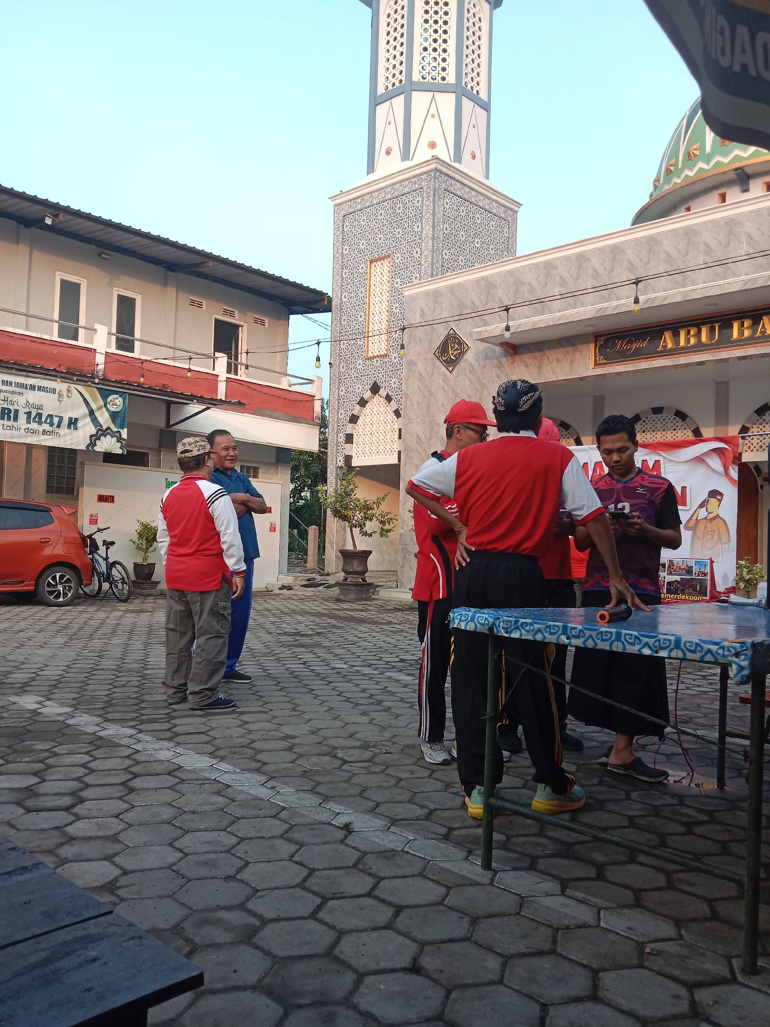 The image depicts an outdoor scene in front of a building with a mosque minaret and dome, located in Madiun, Indonesia. A group of men are gathered, some standing and talking, while others appear to be interacting with a table set up outdoors. One man in a red and white shirt is looking at his phone, and another man in a dark t-shirt is also holding a phone. A table covered with a patterned cloth is in the foreground. The ground is paved with hexagonal tiles. In the background, there's an orange car and a bicycle parked near a building. A banner with text and graphics is visible on the building facade. The lighting suggests it is either early morning or late afternoon.