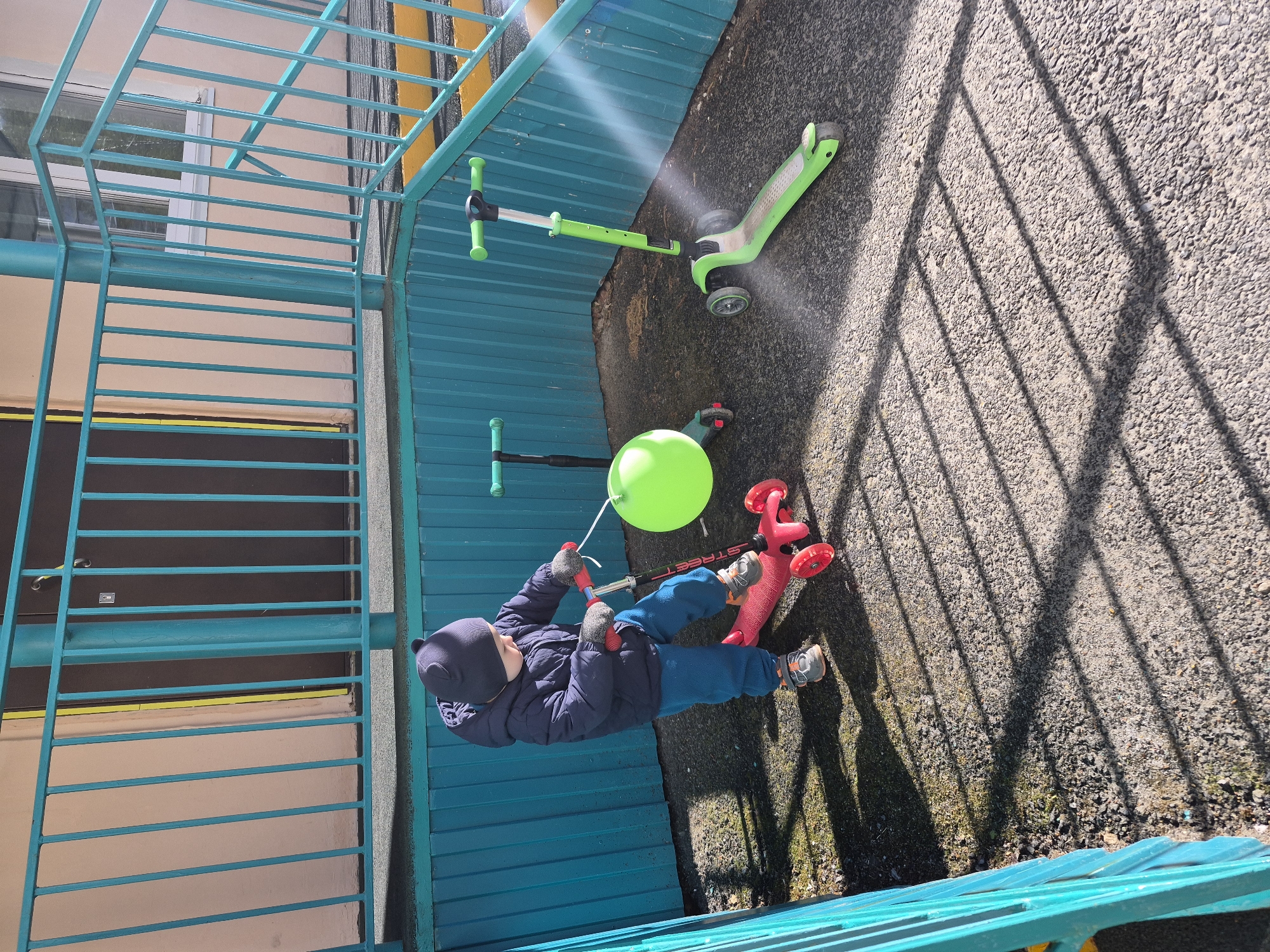 A child in a navy jacket, blue pants, and a navy hat holds a green balloon while standing next to a red scooter on a paved surface. The location is Tobolsk, Russia. Another green scooter is nearby. A metal structure with blue horizontal bars and green vertical posts surrounds the area. The surface casts shadows from the metal structure.