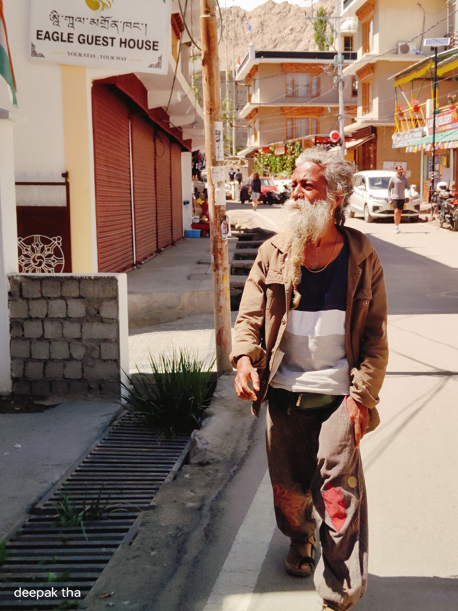 An elderly man with a long gray beard and hair walks down a street in Leh, India. He is wearing a brown jacket over a striped t-shirt and loose-fitting brown pants. A gold chain is visible around his neck. He is wearing sandals. The street is paved and there are buildings on either side. One building has a sign that reads "Eagle Guest House." The man is walking towards the right side of the frame. The background shows more buildings and some mountains. The sunlight casts shadows on the street.