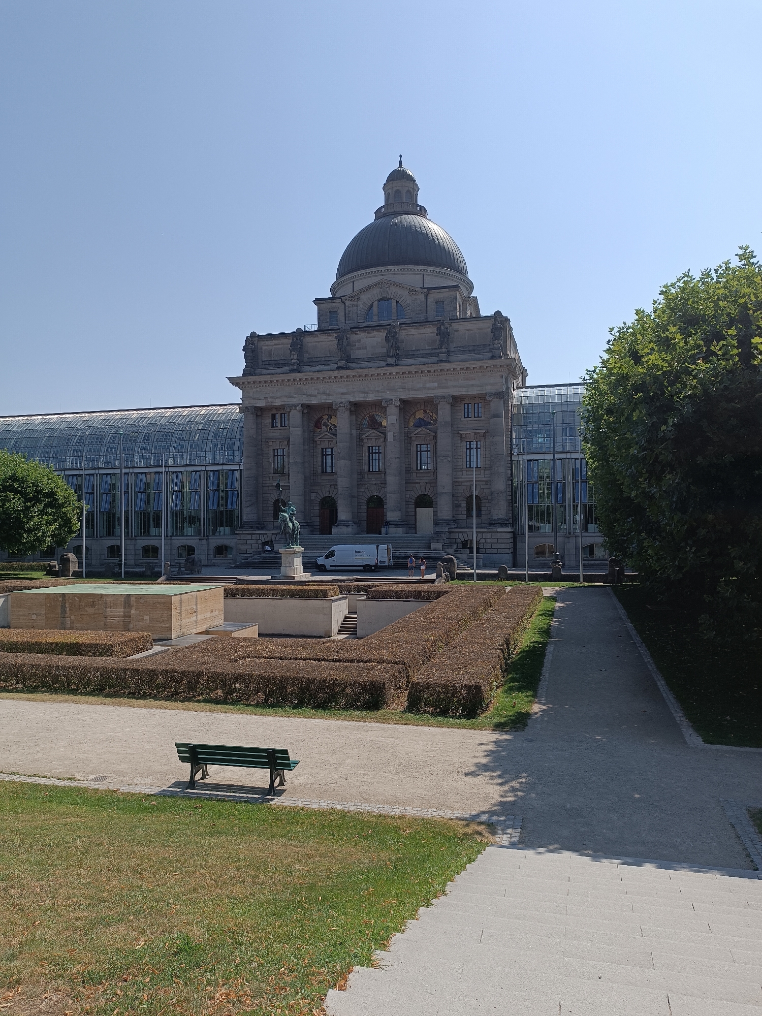 The image depicts a sunny day in front of the Bavarian State Chancellery in Munich, Germany. The grand stone building features a prominent dome and classical architectural elements, with a modern glass structure attached to its side. In the foreground, there is a manicured garden with trimmed hedges and a water feature. A green park bench sits on a gravel path, casting a shadow on the ground. Further back, a statue of a man on horseback stands before the building's entrance, and a white van is parked nearby. The sky is clear and blue, with lush green trees bordering the scene.