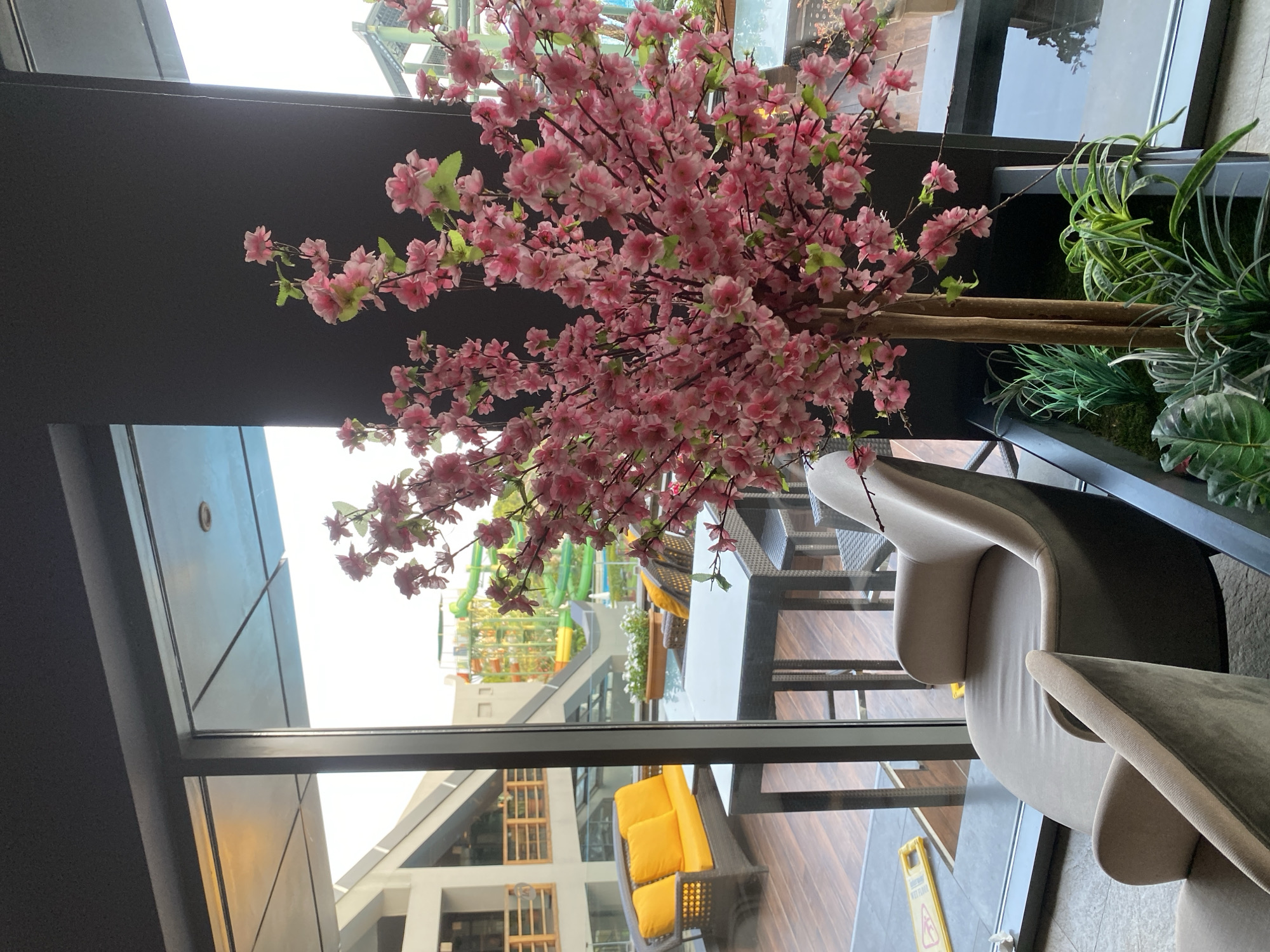 The image captures an indoor seating area with a large window overlooking an outdoor space. In the foreground, a faux cherry blossom tree with pink flowers is prominent, positioned in a planter box with various green plants. To the left of the tree, two plush armchairs are visible. Through the window, an outdoor patio is discernible, featuring a rattan sofa set with bright yellow cushions and a glass-top dining table with chairs. Beyond the patio, a water park with colorful slides can be seen, suggesting the location is likely within a resort or entertainment complex. The overall environment appears to be a modern, stylish seating area designed for relaxation and enjoyment, potentially within a hotel, restaurant, or similar establishment in Aksu, Turkey.