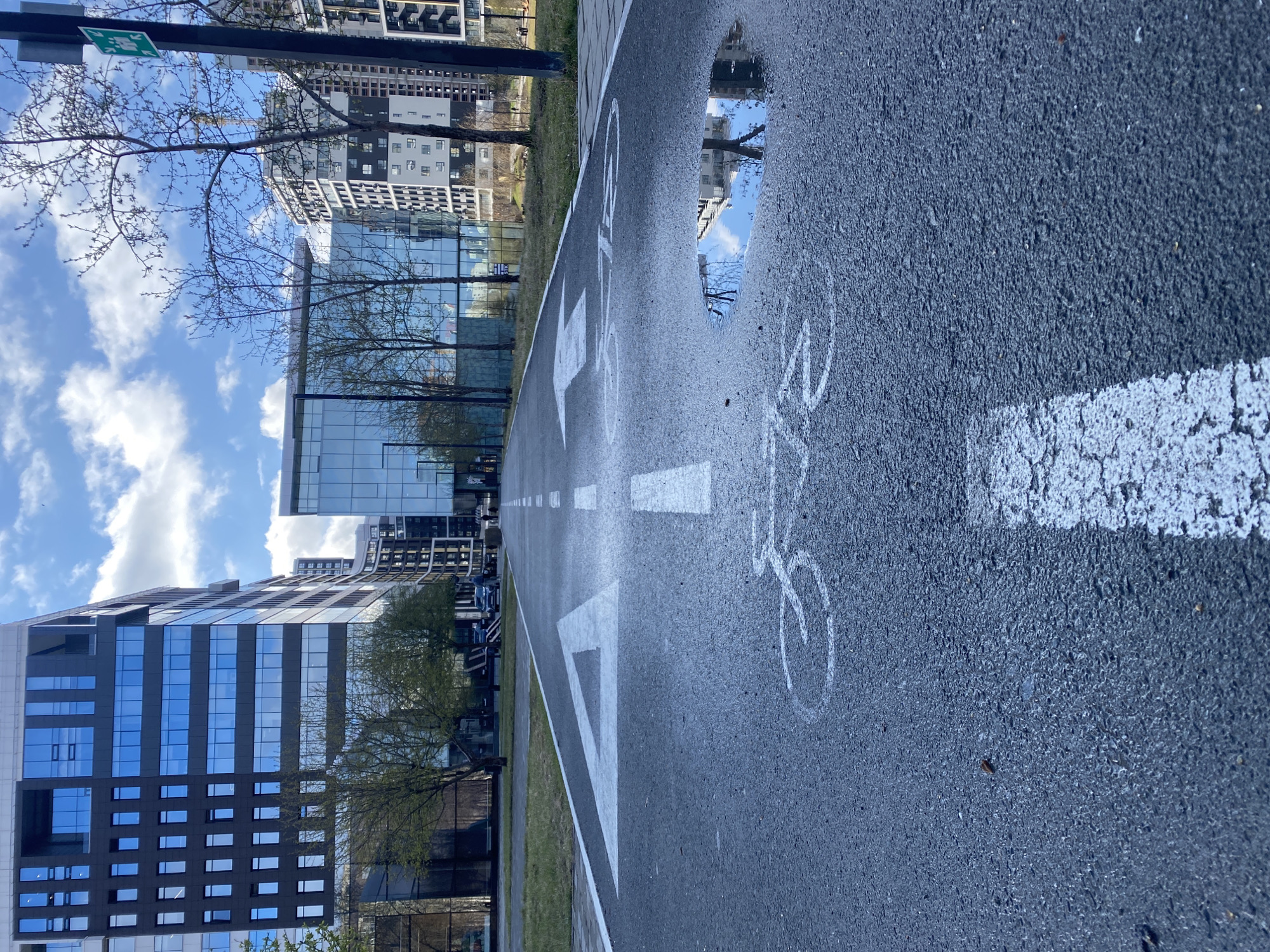 The image captures a modern urban landscape in Yekaterinburg, Russia, viewed along a dedicated asphalt bicycle path under a bright sky. The path, visible in the foreground and extending into the distance, is marked with clear white lines, directional arrows, and multiple bicycle symbols.
On the right side of the path, a large puddle of water reflects the blue sky and the surrounding architecture, suggesting recent rain. The reflection in the puddle also clearly shows building facades and parts of the sky.
Lining the bicycle path are various buildings. To the left, several contemporary multi-story structures feature reflective glass facades and dark panels, their surfaces mirroring the clouds and other buildings. Further in the background, more residential or office buildings with various architectural styles are visible. Scattered trees with mostly bare branches, and some showing nascent green leaves, dot the landscape between the path and the buildings.
The sky overhead is predominantly blue with scattered white clouds, indicating fair weather despite the recent rain. A partially visible green directional sign with a white arrow pointing right is present on the left side of the upper frame. The scene appears calm and well-maintained, characteristic of a modern city environment during daytime.