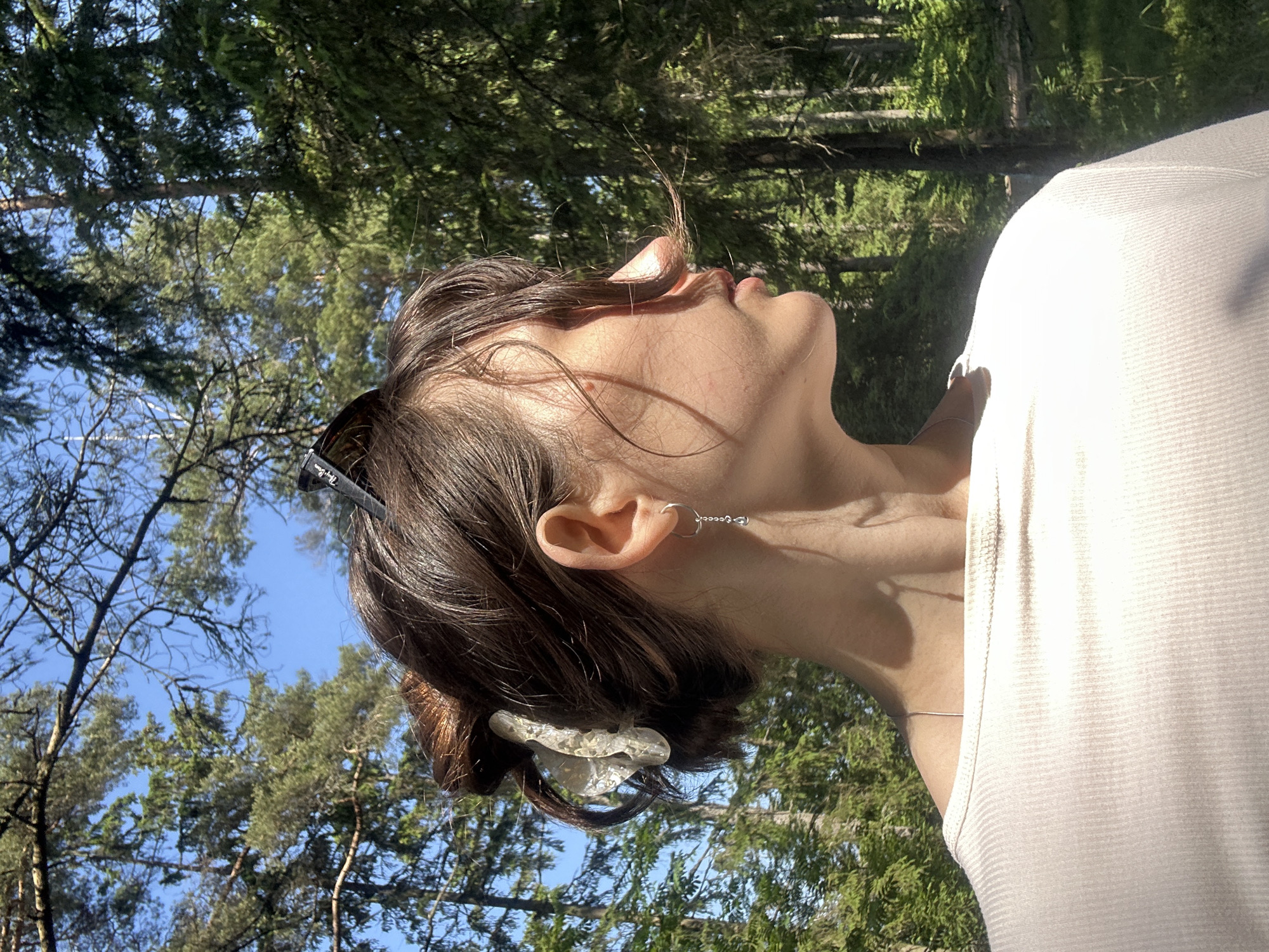 The image shows a young woman from the chest up, in profile, facing right. She has her hair pulled back in a bun and is wearing a white ribbed top. She has a silver earring in her left ear and sunglasses perched on top of her head. The background is a forest with tall trees and a clear blue sky. The lighting suggests it is daytime. The image appears to have been taken outdoors in a natural setting. No specific details about Shepelyovo, Russia are visible in the image.