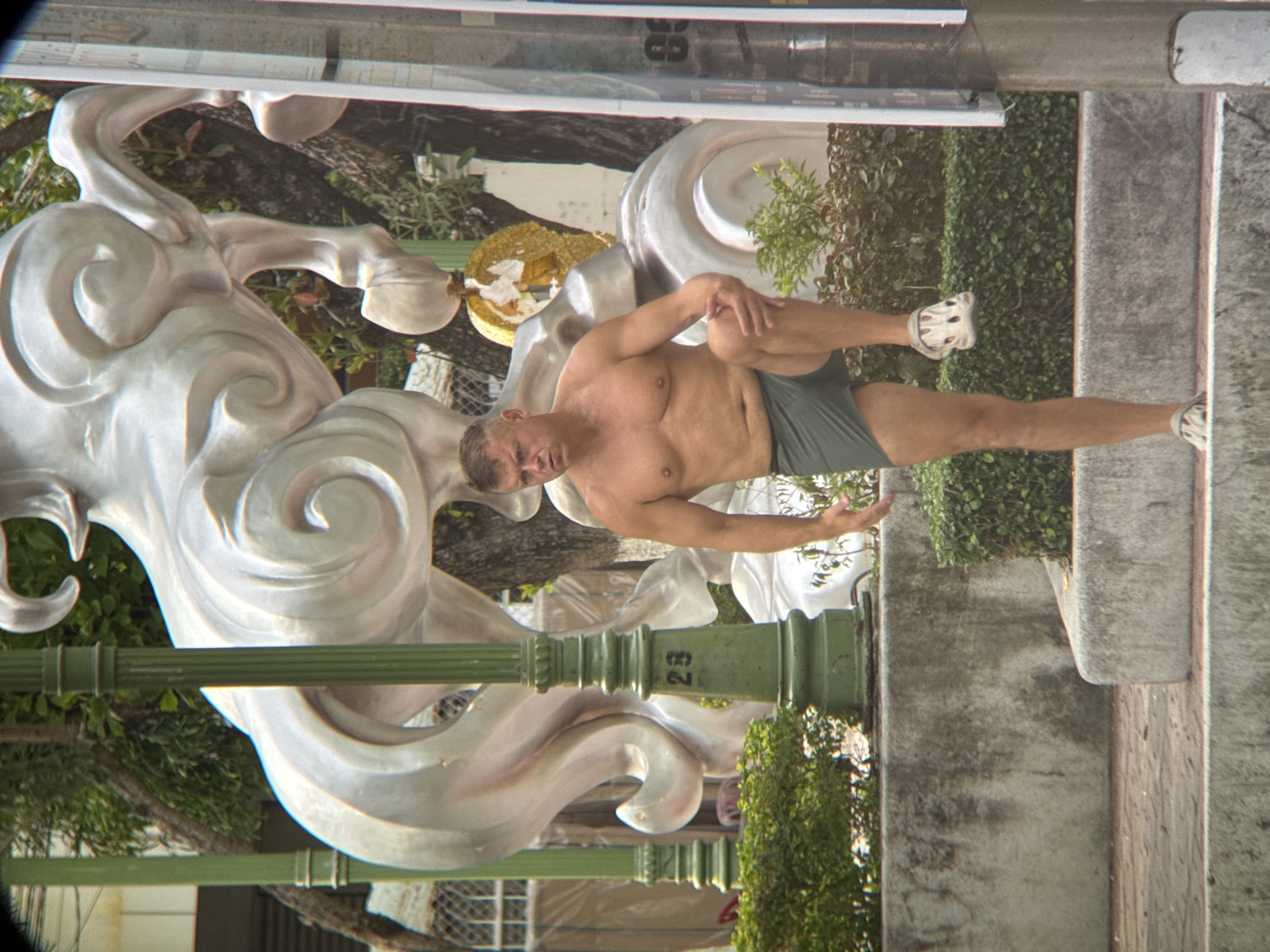 A shirtless, middle-aged man with short brown hair and a light complexion is standing outdoors in front of a large, silver sculpture of a horse, which is partially obscured by trees and foliage. The man is wearing dark green shorts and white slip-on shoes. He is looking to his left and has his right leg bent at the knee, with his foot resting on a lower surface. His left leg is extended, and he is holding his left hand on his left knee. In the background, there are green lampposts, a wall, and more greenery. The sculpture appears to be a modern, abstract representation of a horse, with flowing lines and curves. The scene is set in an urban environment, possibly a park or public space. The man's posture suggests he might be stretching or posing. The presence of the large sculpture indicates that this is likely an area of artistic or cultural interest. The context of Nha Trang, Vietnam, suggests a warm, possibly tropical climate.