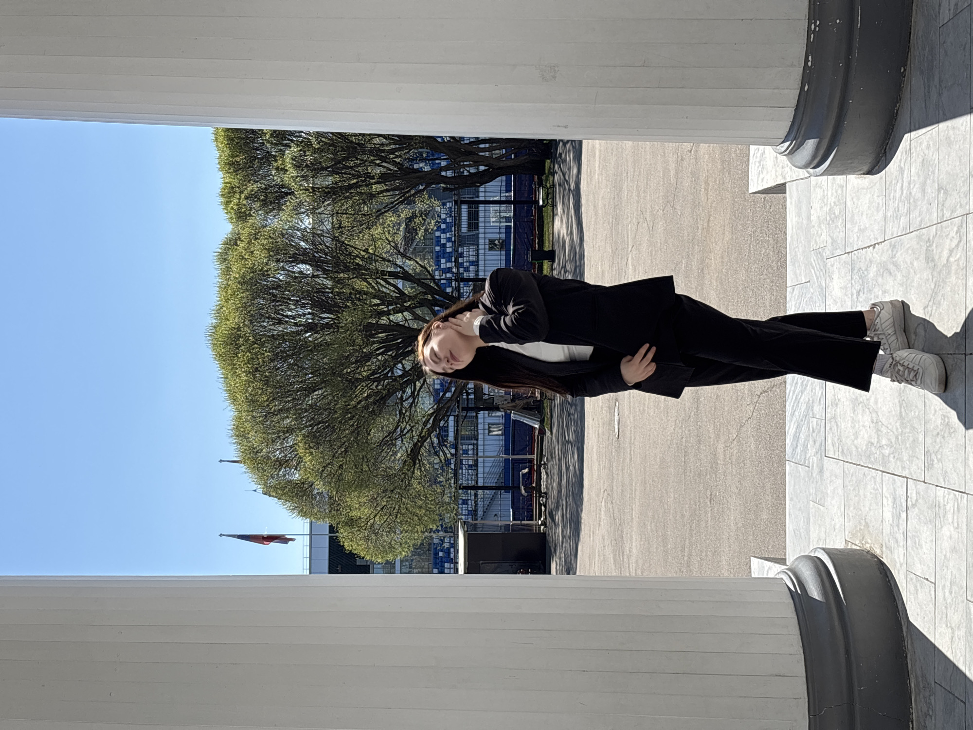 A young woman stands between two large white columns, likely made of marble, under a clear blue sky. She is wearing a dark business suit, a white shirt, and white sneakers. The woman is leaning slightly and appears to have her hands behind her neck. Beyond the columns, trees with light green leaves can be seen. In the background, there is a building with blue and white checkered panels, a black truck, and two flags flying in the distance. The ground is paved with light grey stone. Based on the visual cues of the architecture and landscape, the location is likely in Perm, Russia.