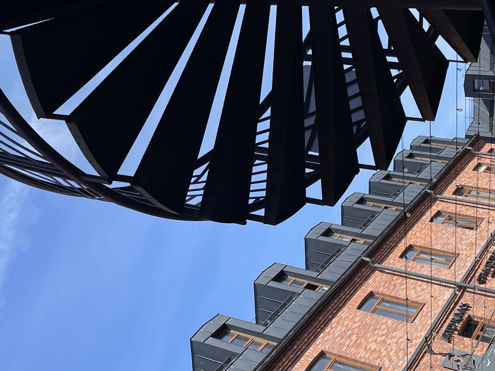 A low-angle shot depicts an architectural view in Saint Petersburg, Russia. The lower right portion features a red brick building with multiple windows and dark gray, geometric dormers, and the building has a sign in Russian. The upper left portion shows a black, spiraling metal staircase against a clear blue sky. The perspective suggests the photographer is looking up from below the staircase, capturing the contrast between the staircase's modern, industrial design and the building's classic, brick facade.