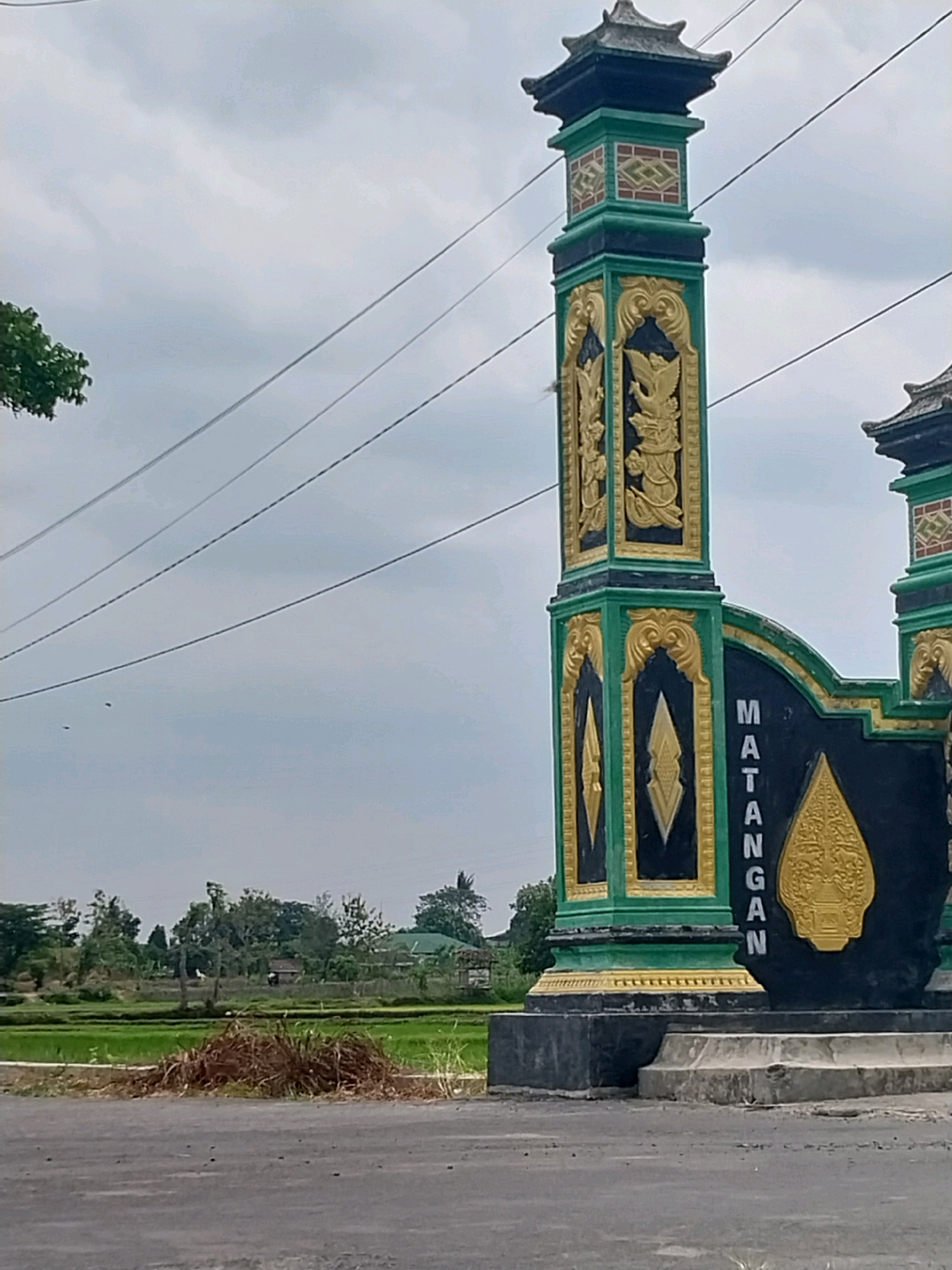 The image shows an ornate, brightly colored gate with the word "MATANGAN" written vertically in black with yellow lettering on a black background. The gate is flanked by a tall, square pillar decorated with green and gold patterns, including what appears to be a stylized bird or mythical creature. The pillar has a tiered roof structure. The gate and pillar are situated near a road, with a backdrop of lush green rice paddies and scattered trees under a cloudy sky. The setting appears to be a rural or semi-rural area. The location is identified as Cucukan, Indonesia.