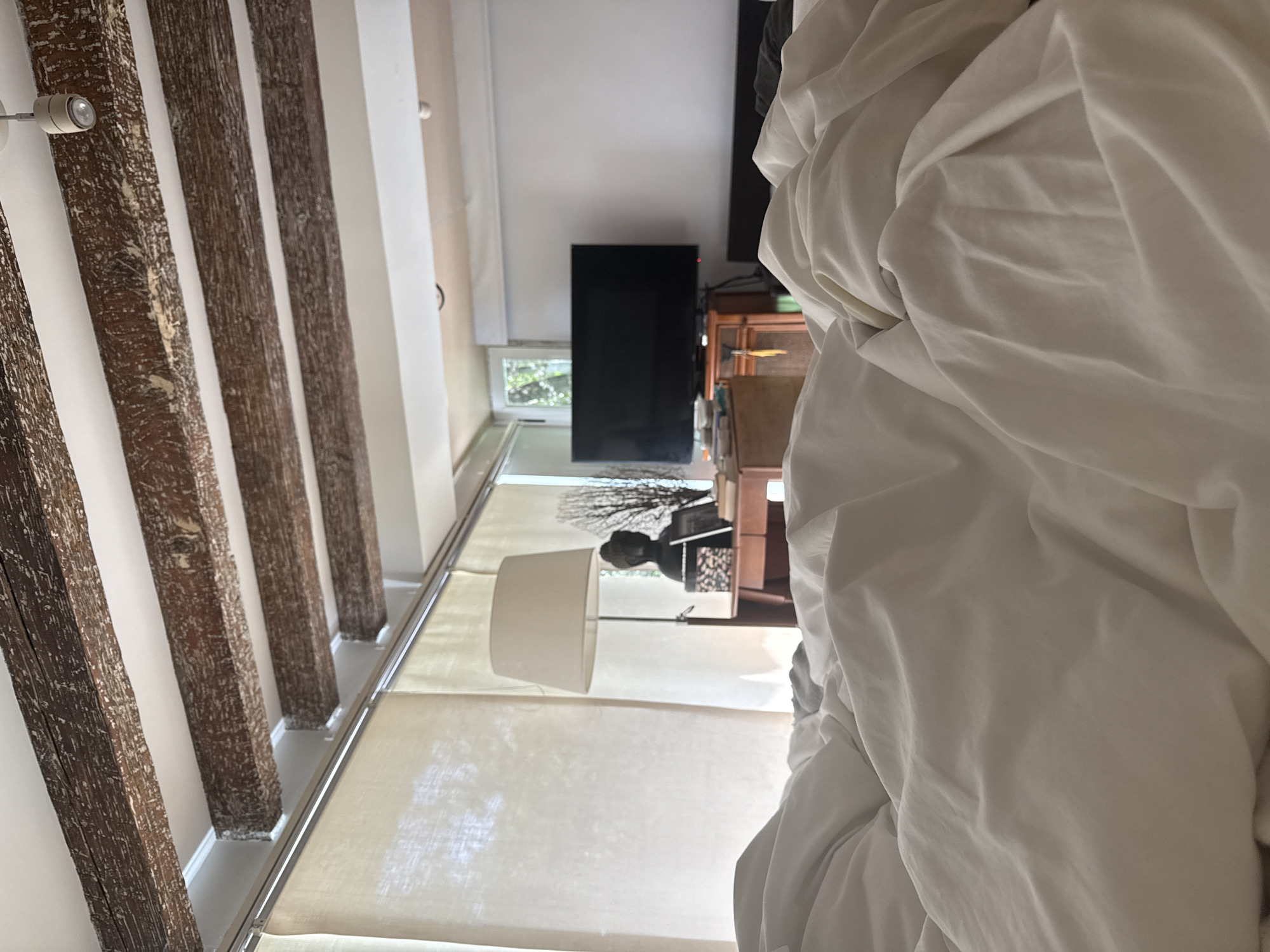 The image depicts an indoor scene, likely a bedroom, with a bed in the foreground covered in rumpled white sheets and duvet. The perspective is from the bed, looking towards the rest of the room. Visible in the background is a large black television screen mounted on a wooden cabinet. To the left of the television, a desk with a lamp and a Buddha statue is present. Behind the desk and lamp, sheer curtains are drawn, revealing a glimpse of greenery outside a window. The ceiling features exposed wooden beams, suggesting a rustic or historical architectural style. The overall ambiance is calm and relaxed, possibly indicating early morning or late evening. The image does not provide any explicit geographical markers to pinpoint its location within Ciudad de México, Mexico.