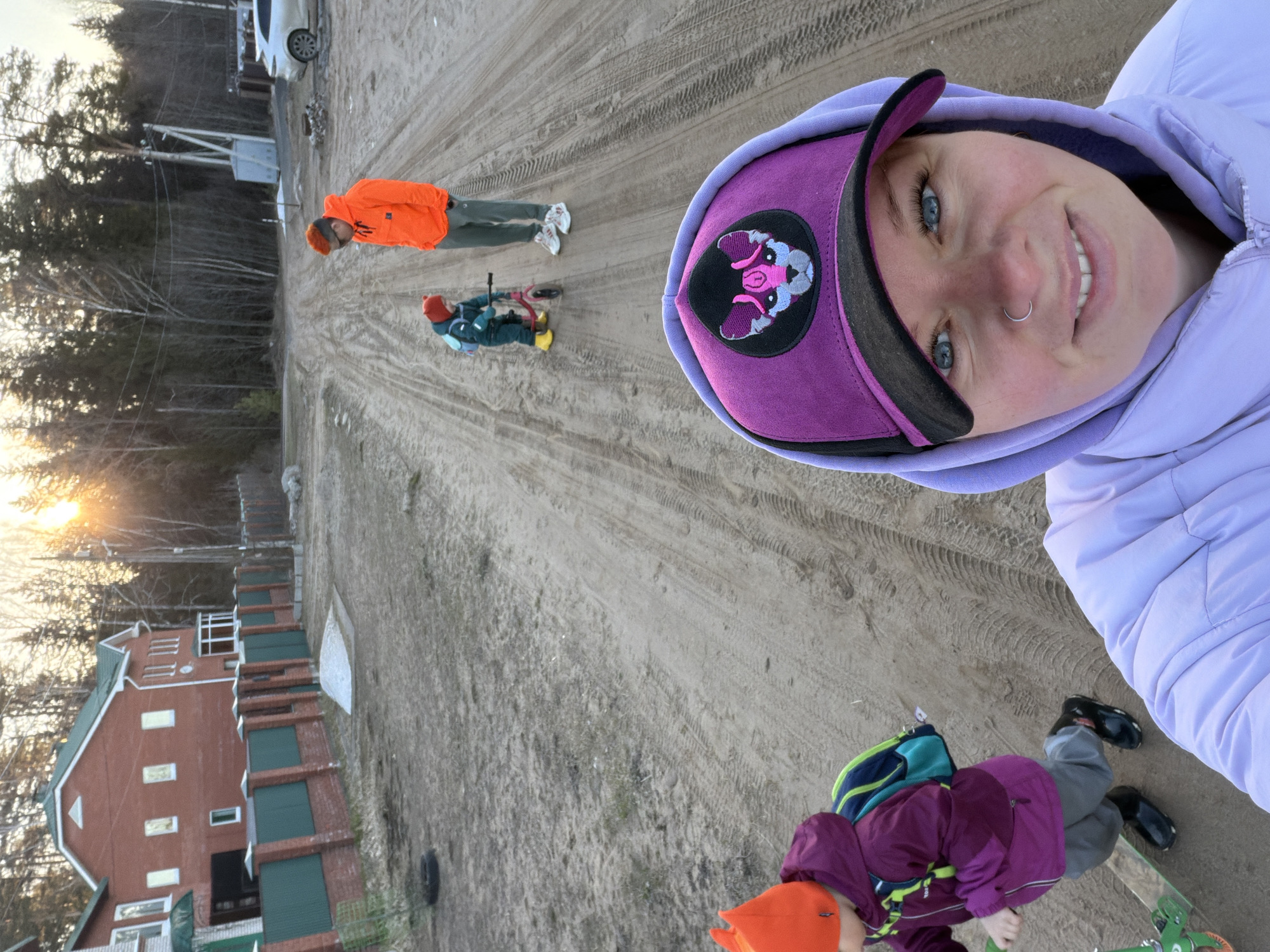 The image, taken from a tilted perspective, captures a scene likely near Chaikovsky, Russia, evidenced by the style of the buildings in the background. The setting appears to be a sandy or dirt-covered area with tire tracks.
In the foreground, a person is taking a selfie, wearing a light purple jacket with a hood and a purple cap with a black brim and a distinctive emblem of a cartoonish pink animal. A second person, lower in the frame, is on a green scooter, wearing a purple jacket, grey pants, and black shoes, with an orange hat.
Further away, an adult in an orange jacket and grey pants stands beside a child on a smaller bicycle.
In the background, there is a house with reddish-brown brick, green shutters, and a green roof. To the right, is a fence that separates the area from a wooded area and another building, a white car is also visible. The lighting suggests it is late afternoon.