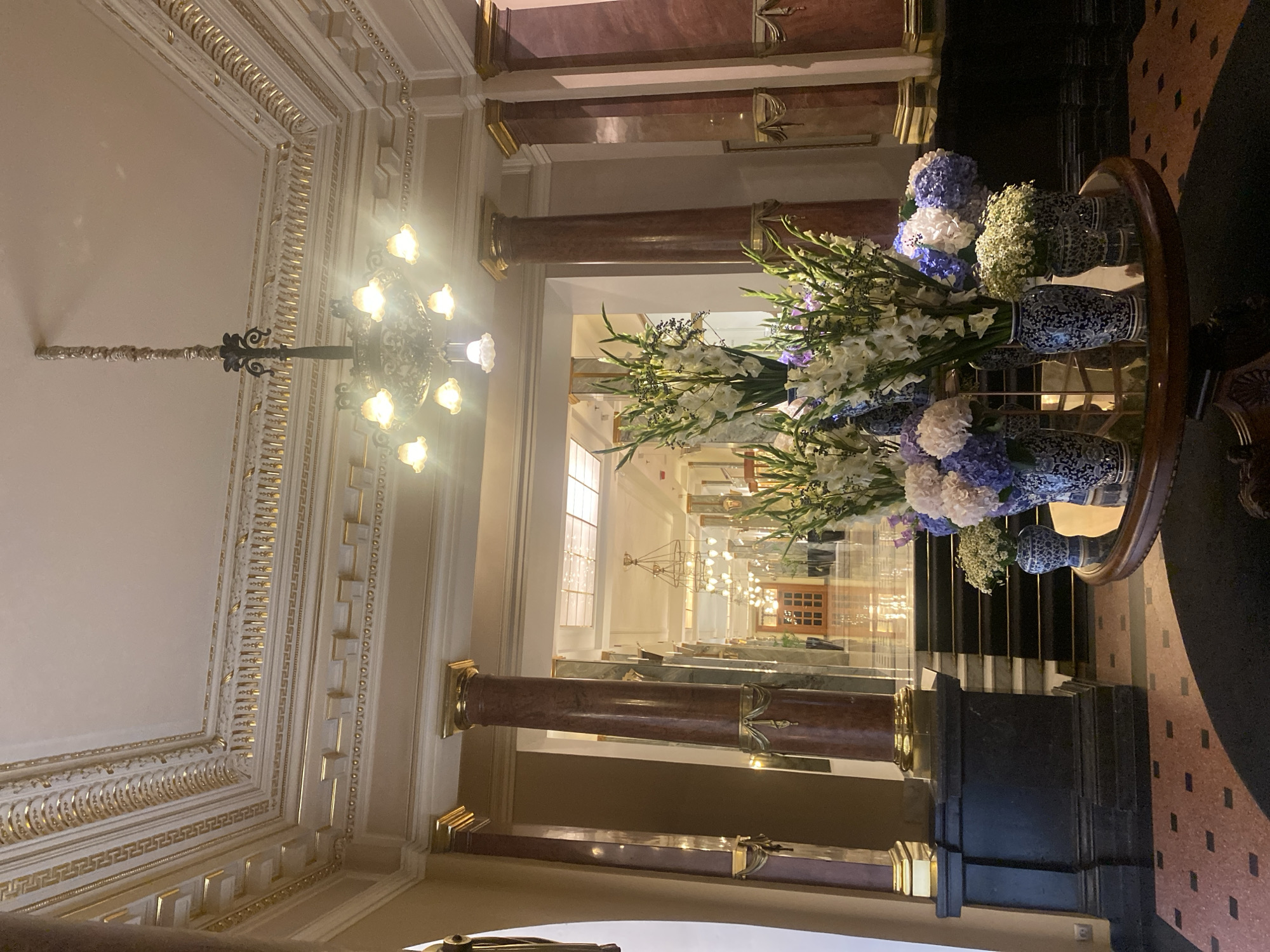 The image depicts an interior scene that appears to be a grand hotel lobby or a similar opulent public space. The scene is captured from a slightly low angle, looking towards a central hallway.
Dominating the foreground is a round, reflective table adorned with several blue and white patterned vases filled with a variety of flowers. These include tall white gladiolus and clusters of blue and white hydrangeas, with some smaller, delicate white blooms. The table itself has an ornate dark wood base.
Behind the table, a wide, carpeted staircase with black treads and risers leads upwards and into a hallway. The hallway is illuminated by a series of chandeliers and natural light filtering in from beyond. The hallway is lined with columns, some of which are made of polished red and green marble, topped with gold accents. The floor in the hallway appears to be highly polished, reflecting the light.
The ceiling is a prominent feature, showcasing intricate gold detailing and ornate molding in a classical style. A large, multi-branched chandelier with glowing lights hangs from the ceiling, adding to the luxurious atmosphere.
The walls are a neutral color, with some areas featuring marble paneling. The overall impression is one of grandeur and sophistication, with an emphasis on rich materials and decorative elements.
Based on the architectural style, particularly the ornate gold ceiling and classical columns, this scene strongly resembles the interior of the Grand Hotel "Bellevue" in Saint Petersburg, Russia. The hotel, known for its historic architecture and lavish interiors, would feature such a space.