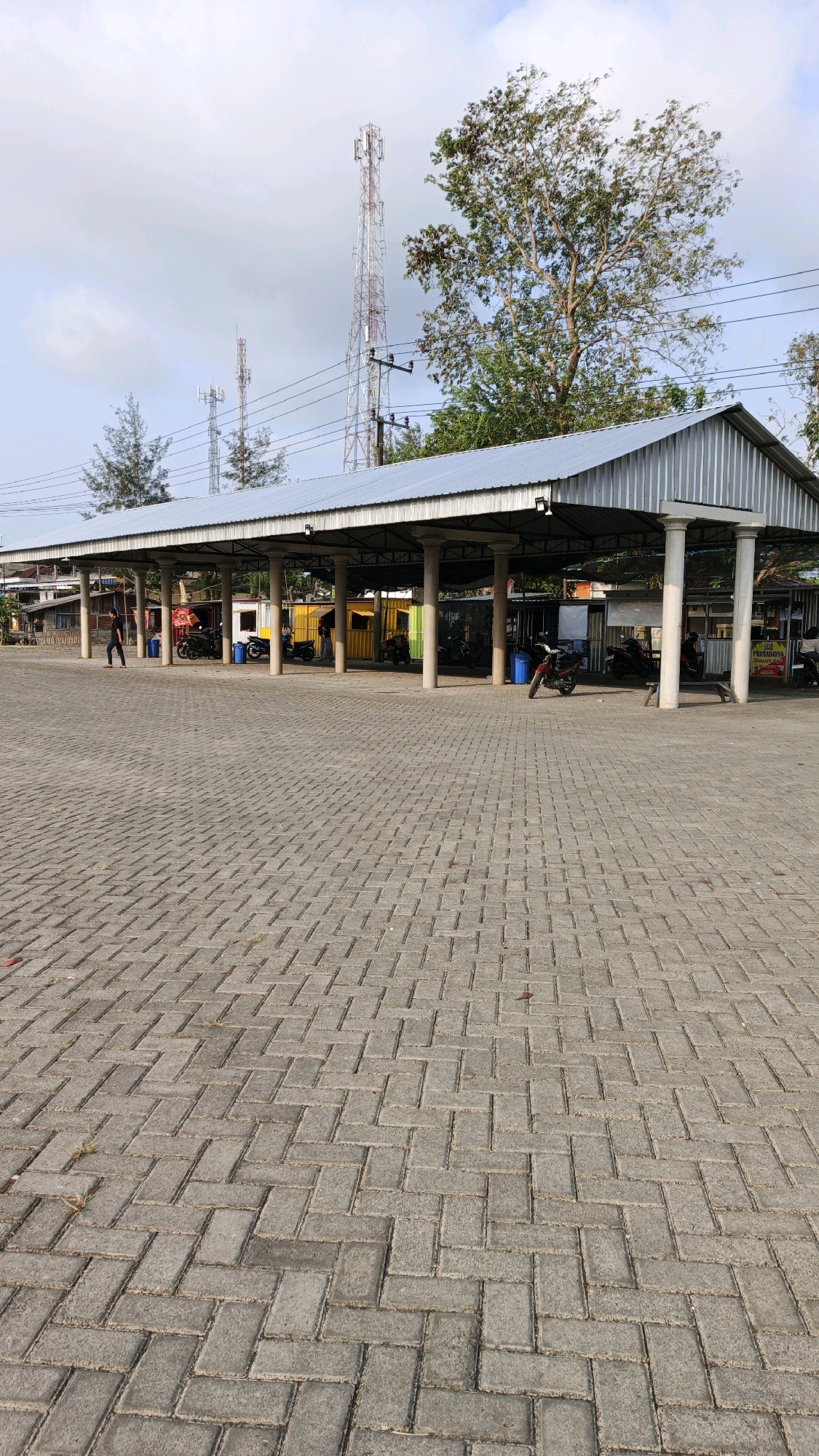 The image depicts a covered parking area with a metal roof supported by numerous white columns. The ground is paved with grey interlocking bricks laid in a herringbone pattern. Several motorcycles are parked under the shelter, and some people are visible in the background, one person walking and others near small stalls or kiosks. In the distance, cellular towers rise against a cloudy sky, and trees are visible. The scene appears to be an open-air marketplace or transportation hub in Tanggulwelahan, Indonesia.