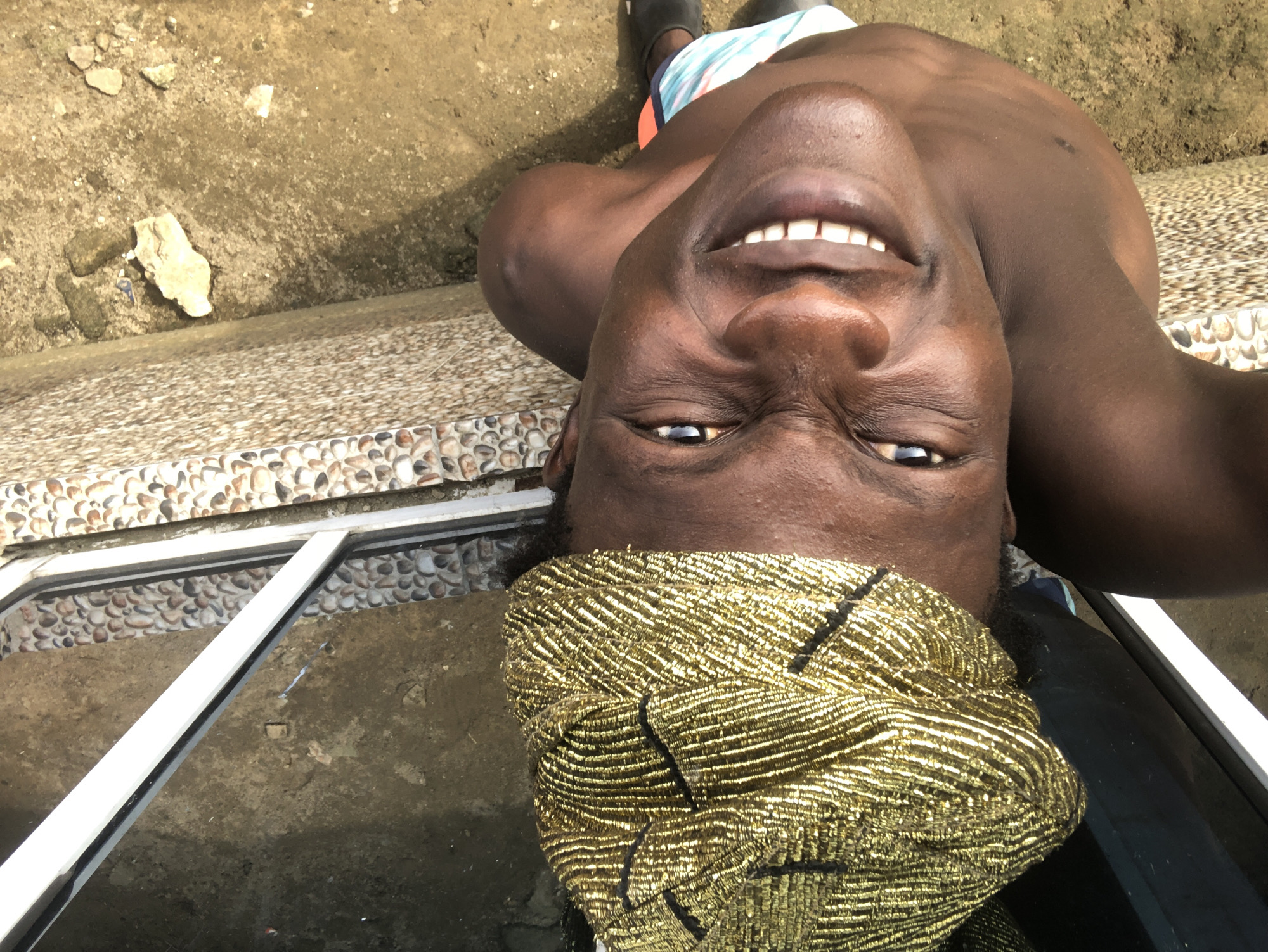 A person wearing a gold-beaded headwrap and colorful swim trunks is lying down outdoors. The person is smiling broadly, with their eyes open. The background consists of a concrete ground, a wall with a pebble mosaic pattern, and what appears to be a window frame. The scene is illuminated by sunlight, casting shadows. The overall impression is of a casual, relaxed moment. The location is not specified in the image, so it cannot be determined if it is in Ado, Nigeria.
