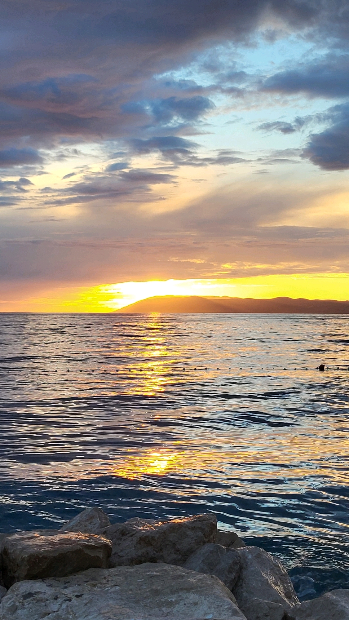 The image depicts a scenic sunset over the sea in Općina Tučepi, Croatia. The sky is filled with a gradient of colors, ranging from deep blues and grays in the upper portion to vibrant oranges and yellows near the horizon. The sun, a bright orb of yellow light, is setting behind a distant mountain range, casting a shimmering reflection on the rippling surface of the water. A thin line of buoys stretches across the sea. In the foreground, large, light-colored rocks are visible, some partially submerged in the water, suggesting a coastal or beach setting. There are no people or animals visible in the scene.