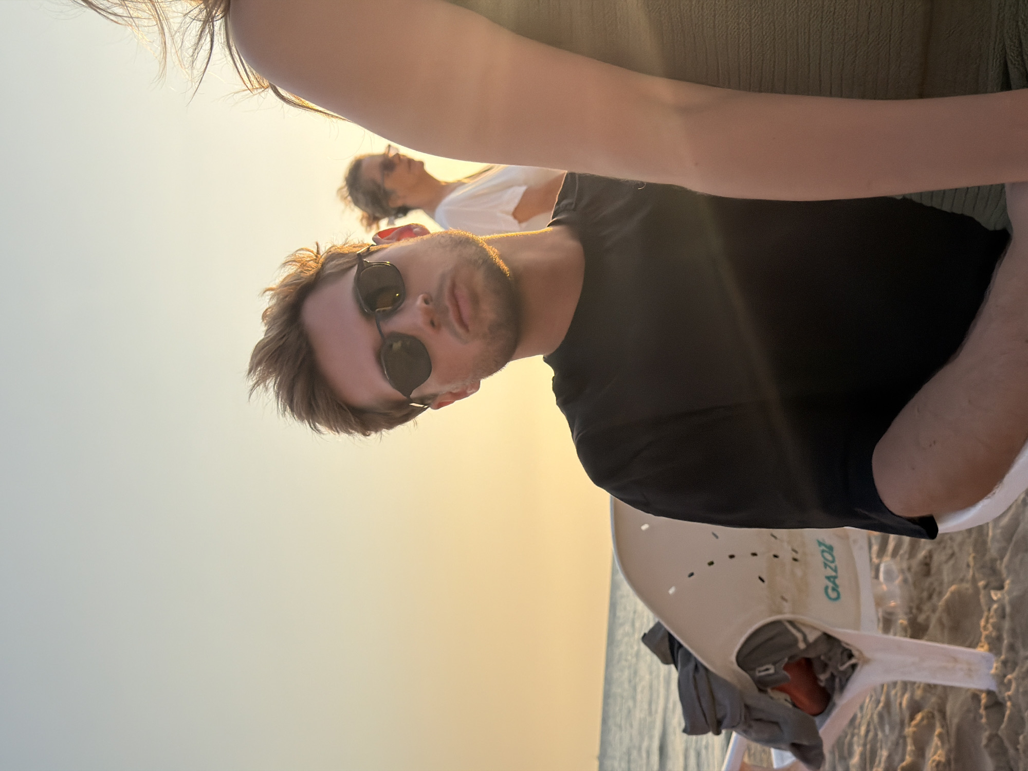 The image depicts a beach scene in Tel-Aviv, Israel, during the golden hour, characterized by warm, low-angle sunlight. Three individuals are visible.
In the foreground, a young adult male with light brown, tousled hair and a short beard is positioned slightly left of center, looking directly at the camera. He wears dark, rectangular sunglasses and a black t-shirt. Strong backlighting creates a golden rim light around his hair, ear, and the left side of his face (viewer's right). He is seated on a white plastic beach chair, which bears the teal word "GAZOI" on its side. A grey garment and an orange object are draped over the back of this chair to his left.
Partially obscuring the right side of the main subject and extending into the right foreground is the right arm and shoulder of another individual, likely female, whose face is not visible. This individual wears a light green or khaki textured garment. Her hair, illuminated by the sun, is visible at the top right corner.
In the mid-ground, behind the main subject and slightly to the right, a third individual, a female with light brown hair pulled back, stands or sits. She wears sunglasses with round frames and a white t-shirt, and is looking towards her right (viewer's left).
The background consists of the calm Mediterranean Sea on the left, meeting a clear sky that transitions from light blue at the top to warm orange-yellow near the horizon line. The ground is sandy.