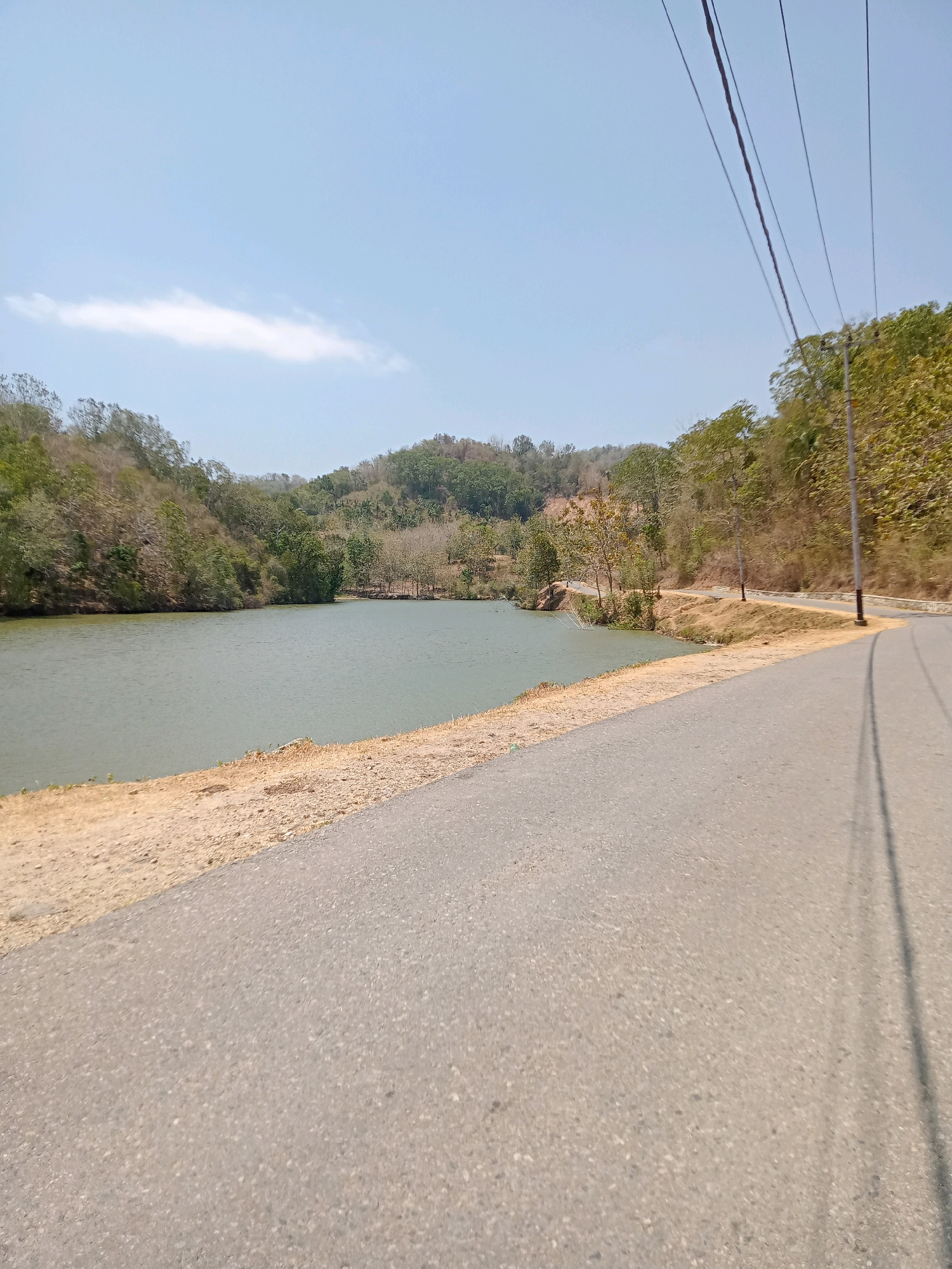 The image shows a paved road curving alongside a calm body of water, likely a lake or reservoir, in Luhu, Indonesia. The water is a muted green color and reflects the clear, pale blue sky above, which features a single wispy white cloud. On the far side of the water, a verdant, tree-covered hillside rises, showing a mix of green foliage and dry, brown branches, suggesting a potentially arid climate or season. The road itself is grey asphalt, with a sandy, dry embankment bordering the water. Overhead, power lines stretch across the frame, supported by a utility pole visible on the right side of the road. The scene appears to be tranquil and natural, with no people or specific structures immediately visible beyond the road and utility infrastructure.