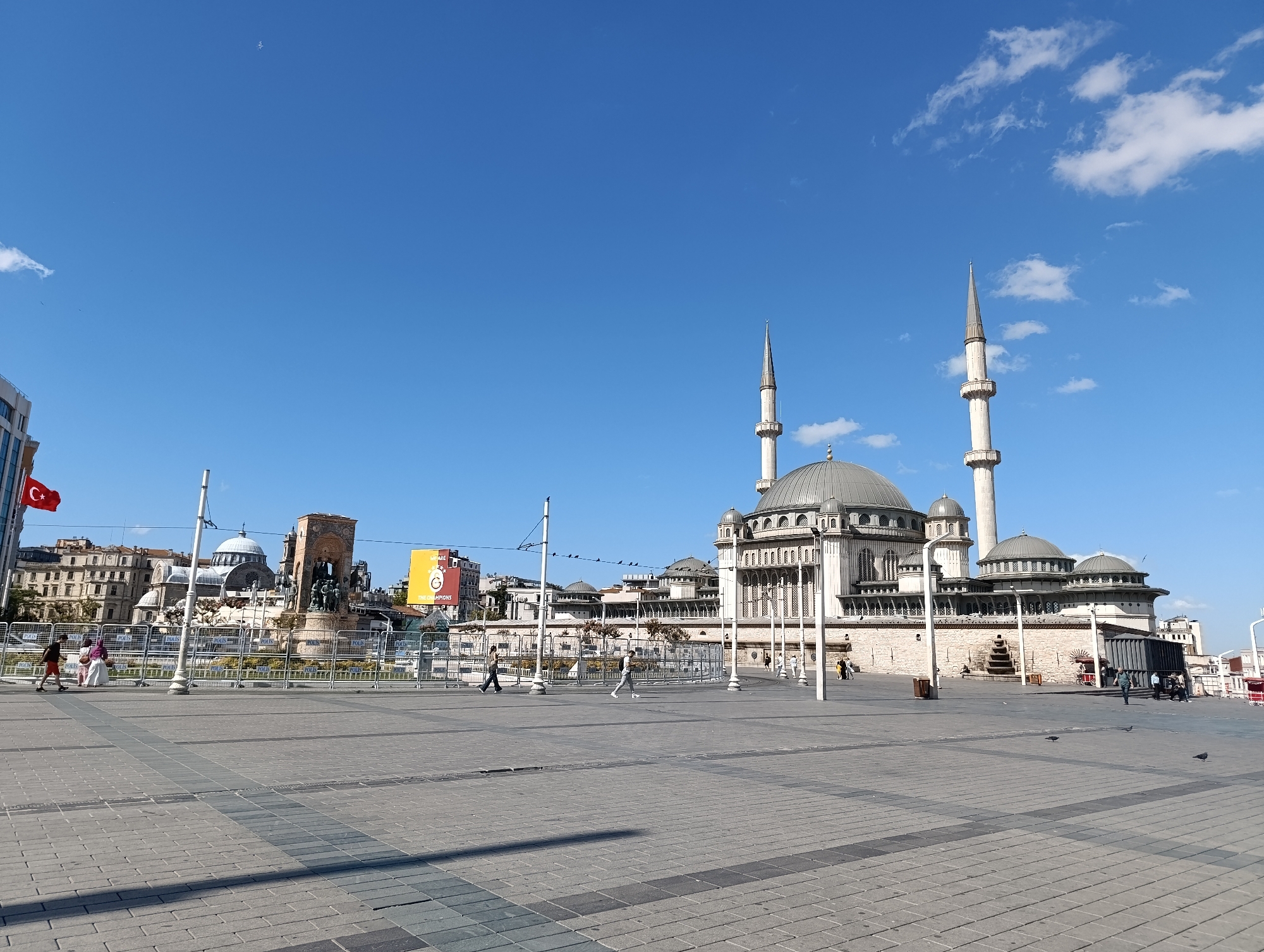 The image depicts the bustling Taksim Square in Beyoğlu, Turkey, on a clear, sunny day. The main focus is the majestic Taksim Mosque, a grand religious structure with its distinctive domes and minarets, standing proudly under a bright blue sky dotted with a few wispy clouds. To the left of the mosque, a prominent monument, likely the Republic Monument, is visible, surrounded by metal barriers. The Turkish flag flutters atop a nearby building.
The square itself is paved with gray brick tiles, showing a clear pattern of individual bricks. Several people are scattered across the expanse of the square, some walking, others standing in small groups. They are casually dressed, suggesting a typical day in a public space. Some are seen near the barriers, observing the sights. In the background, a variety of buildings, including apartments and commercial establishments, line the perimeter of the square. The overall scene conveys a sense of urban activity and cultural significance, with the architectural marvel of the mosque and the historical monument serving as central points of interest.