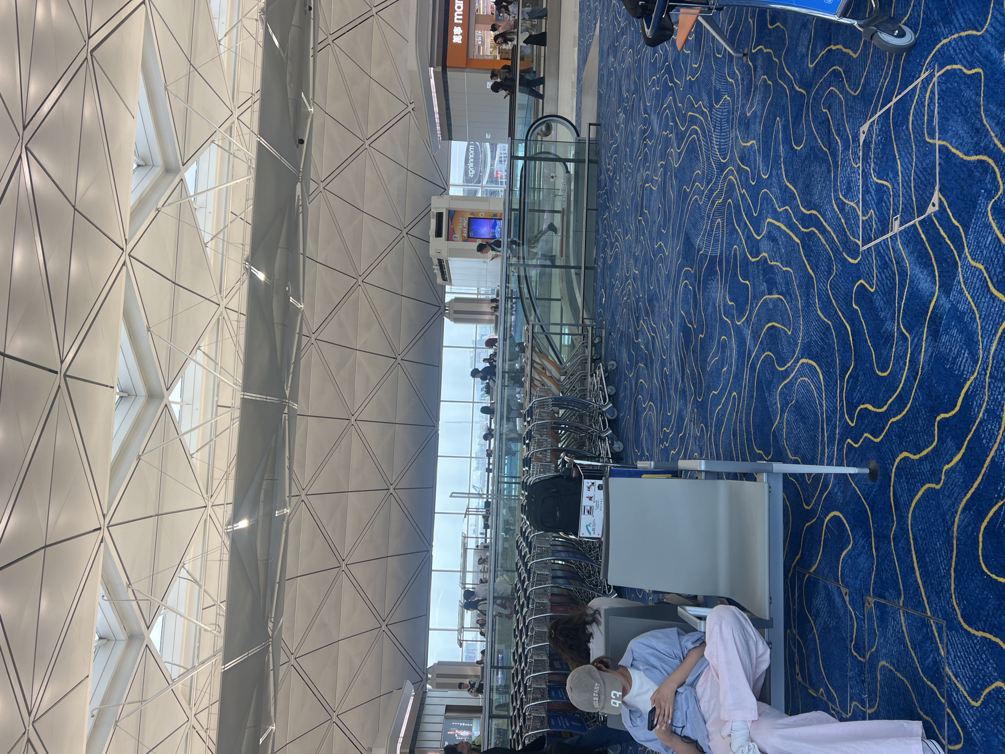 The image depicts the interior of a modern airport terminal, likely Hong Kong International Airport, given its distinctive architectural style, characterized by a large, geometrically patterned ceiling with skylights. In the foreground, a person wearing a blue shirt and pink pants is seated, looking at their phone. Adjacent to this individual is a row of luggage carts. Further into the terminal, an escalator is visible with people moving on it. Shops and departure boards are present in the background, along with large windows offering a view of the tarmac and aircraft. The flooring is a deep blue carpet with a swirling yellow pattern. The environment appears clean and well-lit, suggesting a public transportation hub.