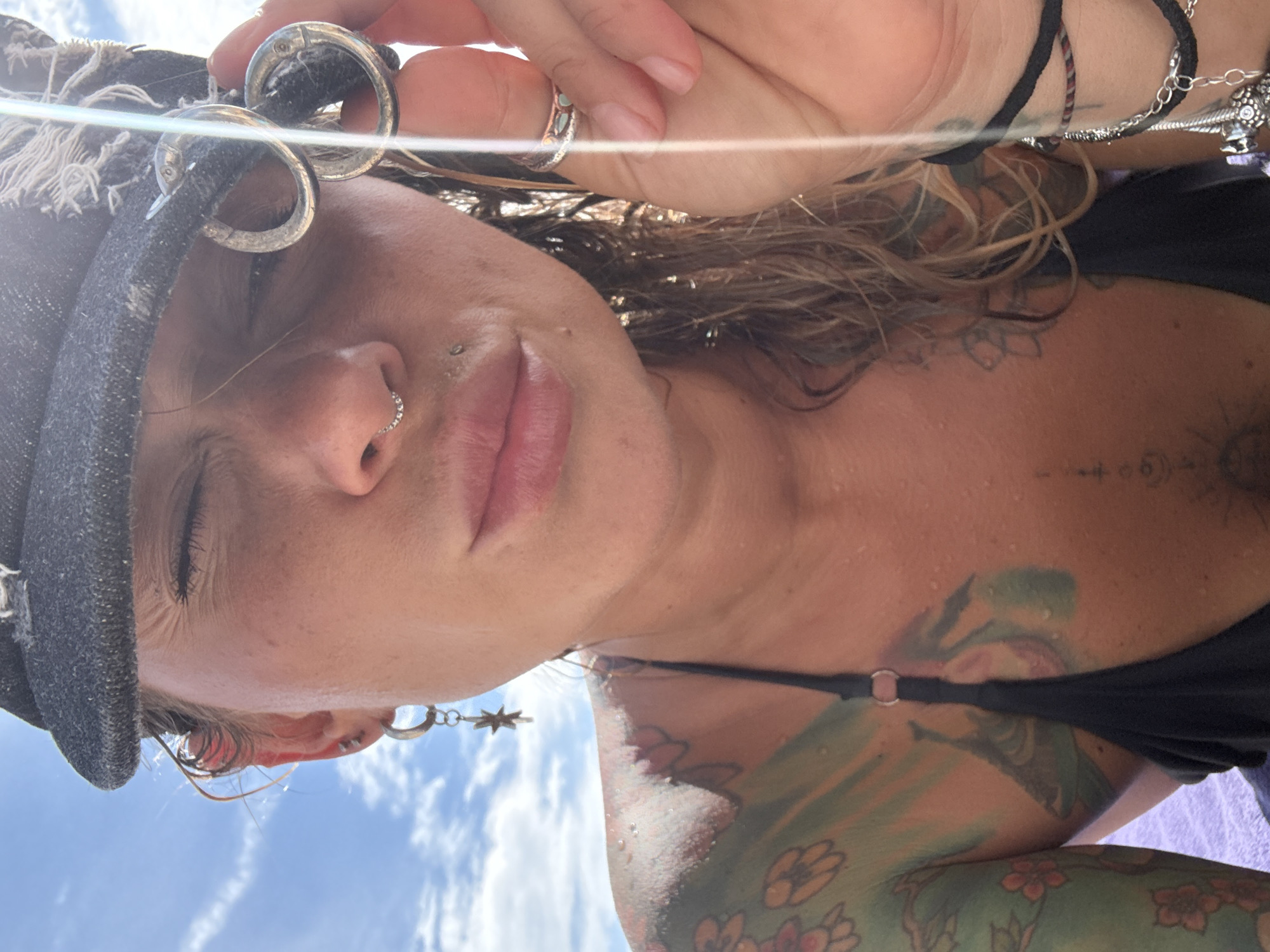 The media file is an extreme close-up, upside-down, selfie-style shot of a person's face and upper torso. The subject is wearing a dark hat with frayed edges and a black bikini top. They have visible piercings on their nose and ear, with a star-shaped earring. The person also has numerous tattoos on their neck, chest, and arm, some of which are floral designs. The skin appears to be slightly dewy, suggesting warm or humid conditions.
In the background, the sky is visible with scattered white clouds, indicating it is daytime. The lighting suggests it could be late morning or early afternoon. The overall tone of the image is casual and relaxed, with the subject's eyes closed and lips slightly parted.
The file is located in Choeng Thale, Thailand. There is no visible text in the image.