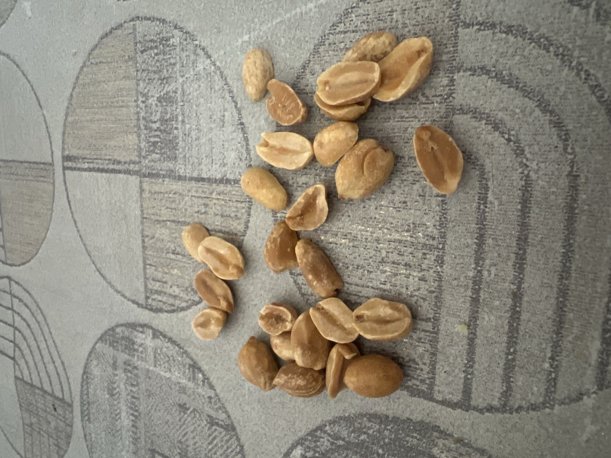 A pile of roasted peanuts is scattered on a textured gray surface. The peanuts are mostly whole, with a few split in half, revealing their inner nut. The surface has a pattern of intersecting circles with a wood-grain texture. The lighting is even, and the peanuts are the primary focus of the image. There are no people or other objects visible.
