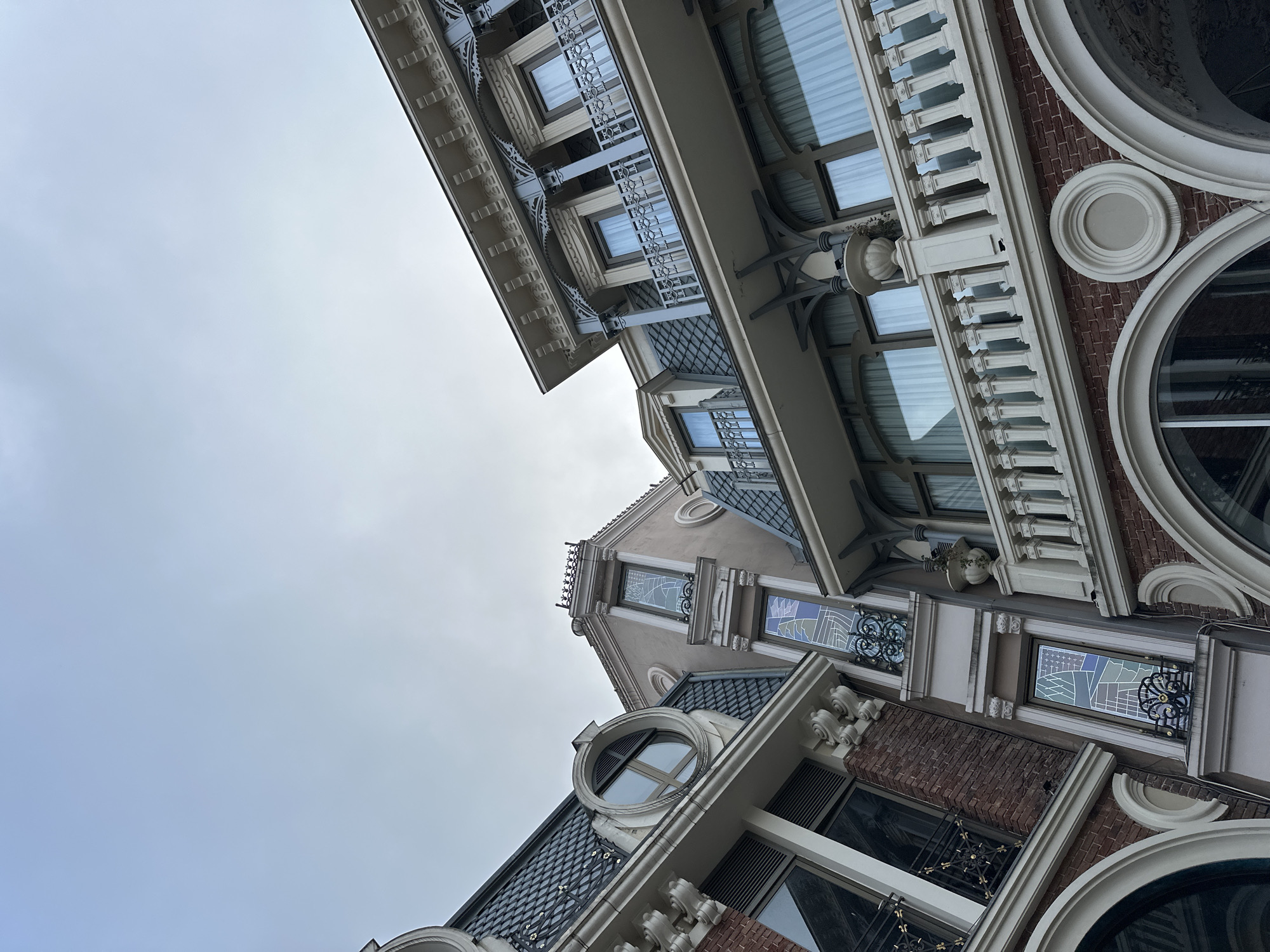 The image shows buildings in Batumi, Georgia from a low angle, looking up. The architecture features ornate facades with decorative elements such as wrought iron, circular windows, brickwork, and light-colored balustrades. The sky is overcast, suggesting diffused light. No people are visible.
