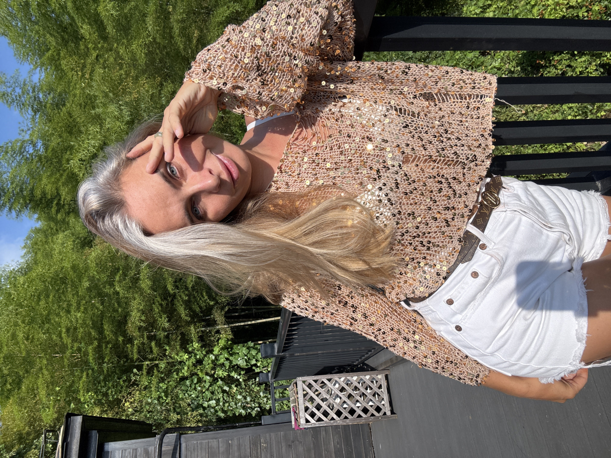 A woman with blonde and graying hair stands on a wooden deck in front of a dense bamboo forest. She is wearing a cropped, loosely knit, sequined top over a white bra and high-waisted white denim shorts. The woman is leaning on a black metal railing, with her right hand touching her forehead and her left hand resting on her thigh. Sunlight casts shadows on her face and clothing. To the left of the woman is a dark wooden building and a wooden lattice trash can. The scene is illuminated by natural sunlight.