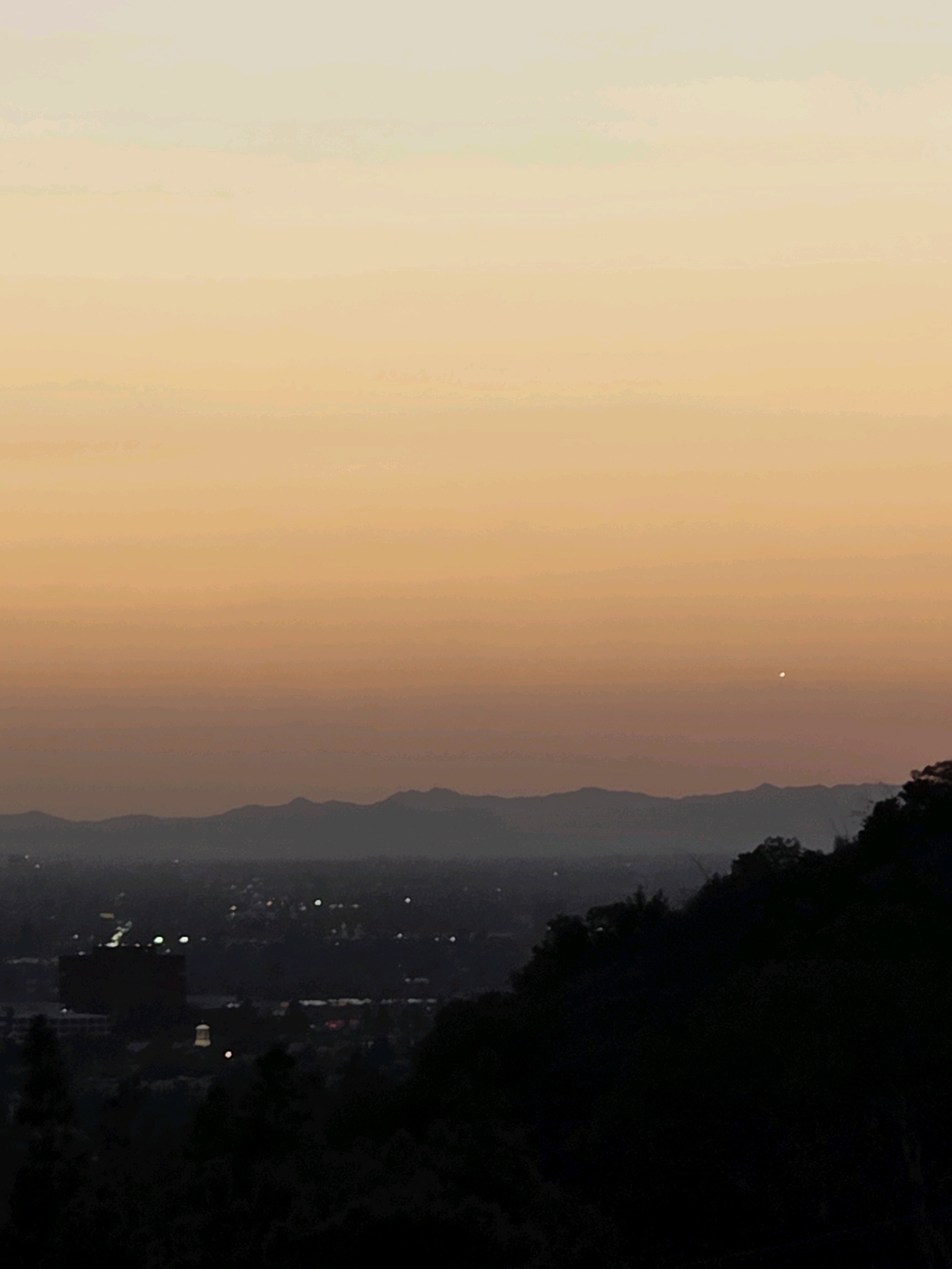 This image captures a panoramic view of Glendale, California, at dusk. The sky transitions from a pale yellow at the top to a soft orange and then a muted pink near the horizon, with a single bright star or planet visible high in the sky. In the distance, a range of hazy, dark mountains forms a silhouette against the fading light. Below the mountains lies the city, with a scattering of lights indicating urban development. One prominent building, a large, dark rectangular structure, stands out among the lights. The foreground is dominated by the dark silhouette of lush green trees and foliage, which are rendered almost black by the low light conditions, obscuring the lower portion of the city. The overall scene suggests a quiet evening over the sprawling landscape of Glendale.