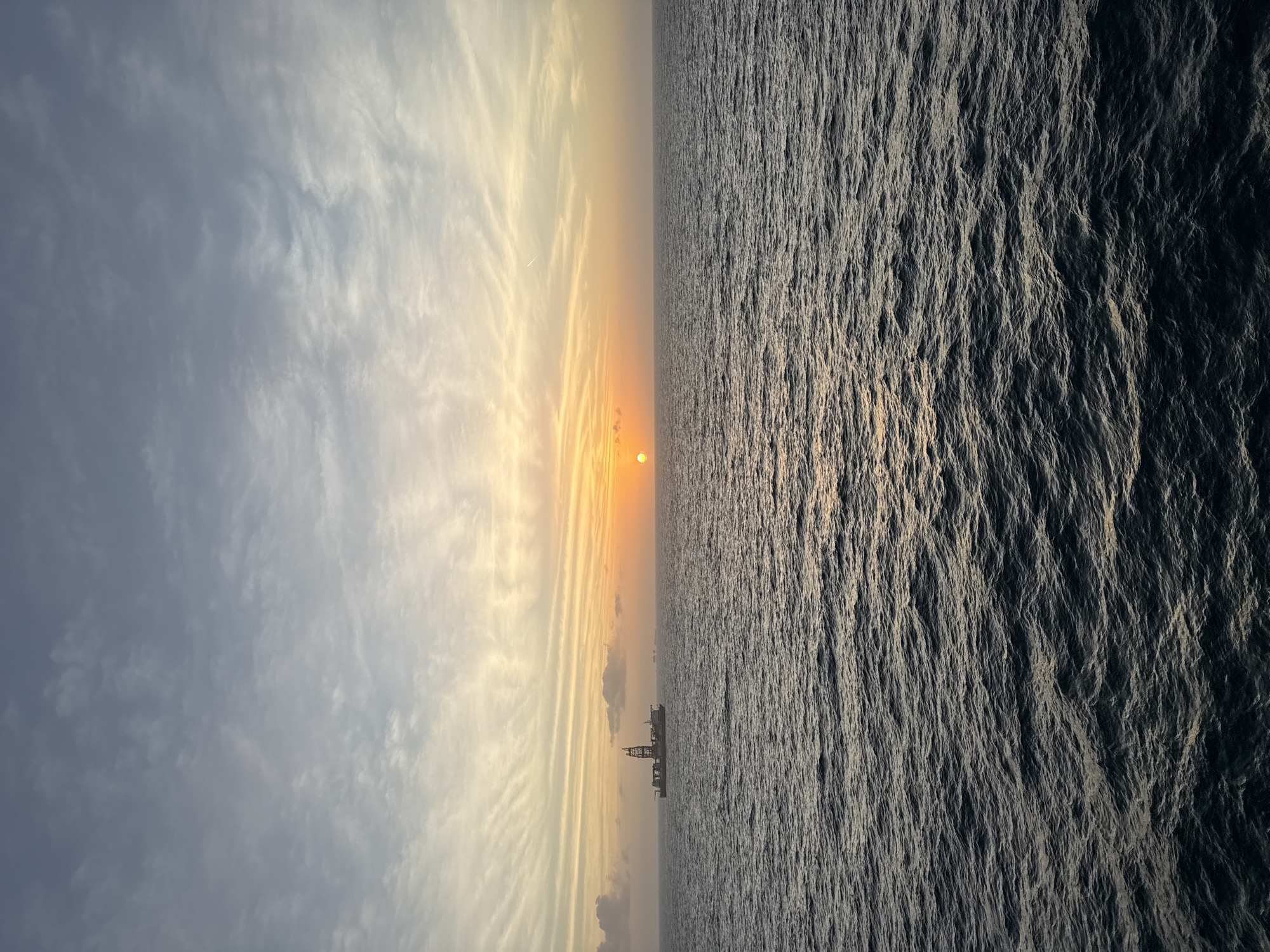 The image shows a sunset over the ocean, with a ship or oil platform visible on the horizon. The sky has wispy clouds, and the water is choppy. The location is unknown.