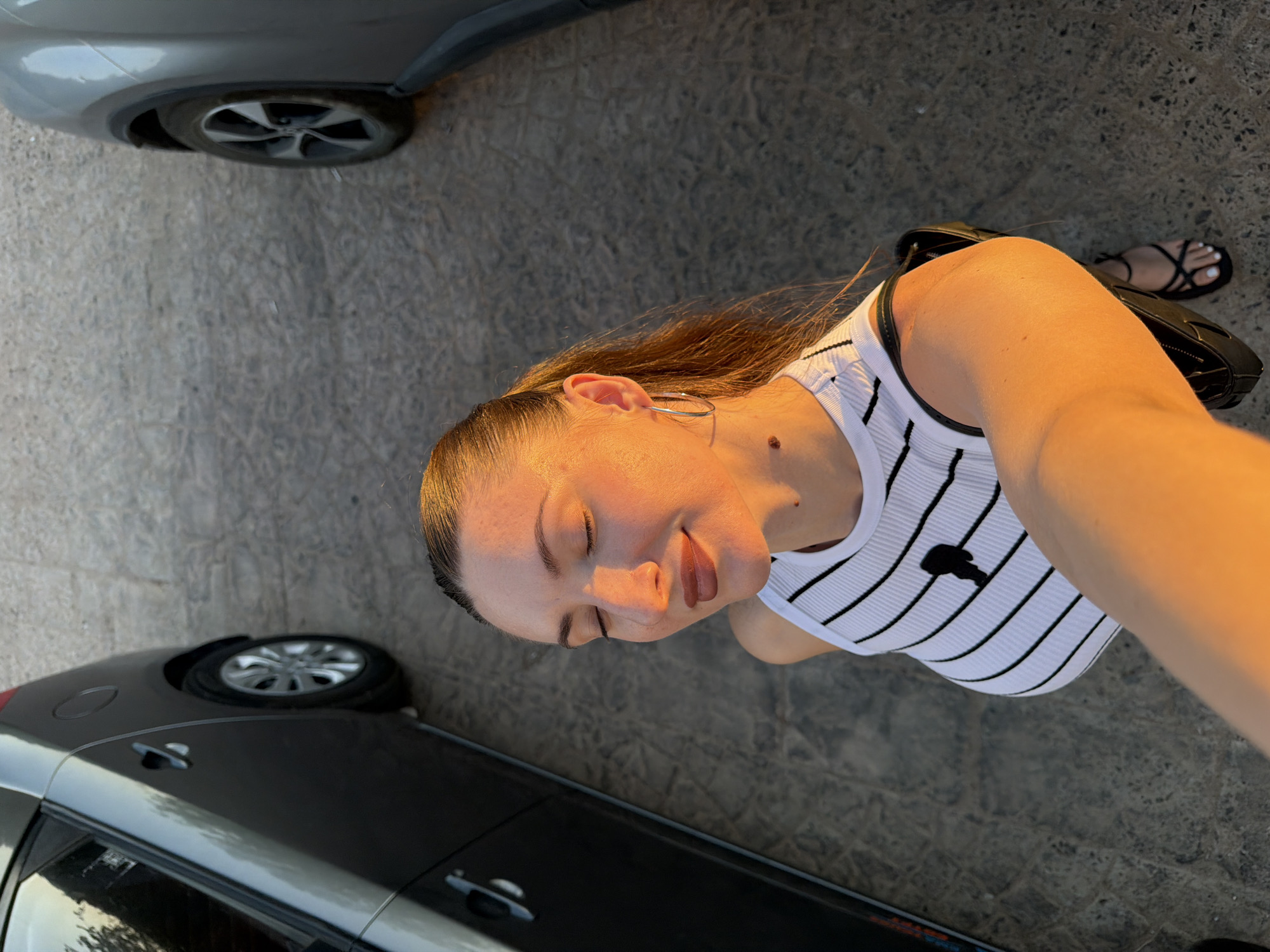An outdoor, eye-level, medium shot captures a young woman taking a selfie in Sharm El Sheikh, Egypt. The woman, with her eyes closed and a slight smile, is bathed in the warm, golden light of a setting sun. She wears a white ribbed tank top with black horizontal stripes and a small black emblem on the chest. Large silver hoop earrings adorn her ears, and her hair is pulled back into a ponytail. A mole is visible on her neck.
She is positioned between two parked cars, one dark gray on the left and another silver on the right. The ground beneath them appears to be a textured, paved surface. The woman is holding a black handbag over her left shoulder, and a black sandal is visible on her right foot. The overall setting suggests an urban or semi-urban environment within Sharm El Sheikh.