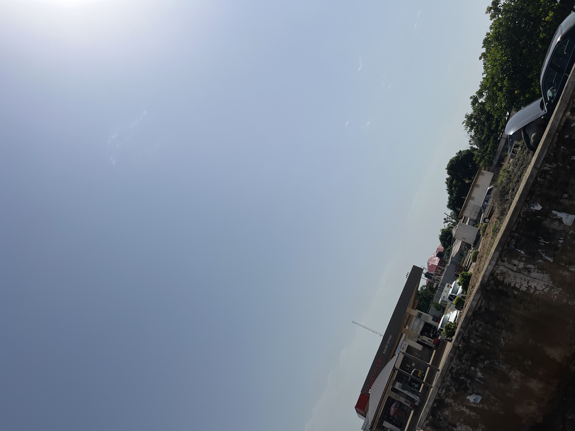 The image depicts a bright, sunny day in Bukuru, Nigeria, with a clear blue sky. In the mid-ground, a TotalEnergies gas station is visible, identified by signage that reads "TotalEnergies BUKURU BYPASS." The gas station has multiple pumps under a canopy. Several cars are parked at or near the station, including a white sedan and a white van. In the foreground, a concrete wall runs horizontally across the lower portion of the frame. To the right of the gas station and beyond the wall, there are lush green trees and some buildings. A dark-colored car with its hood open is parked in the lower right section of the image, with a person partially visible working on it. The scene suggests a typical day at a roadside establishment in Bukuru.