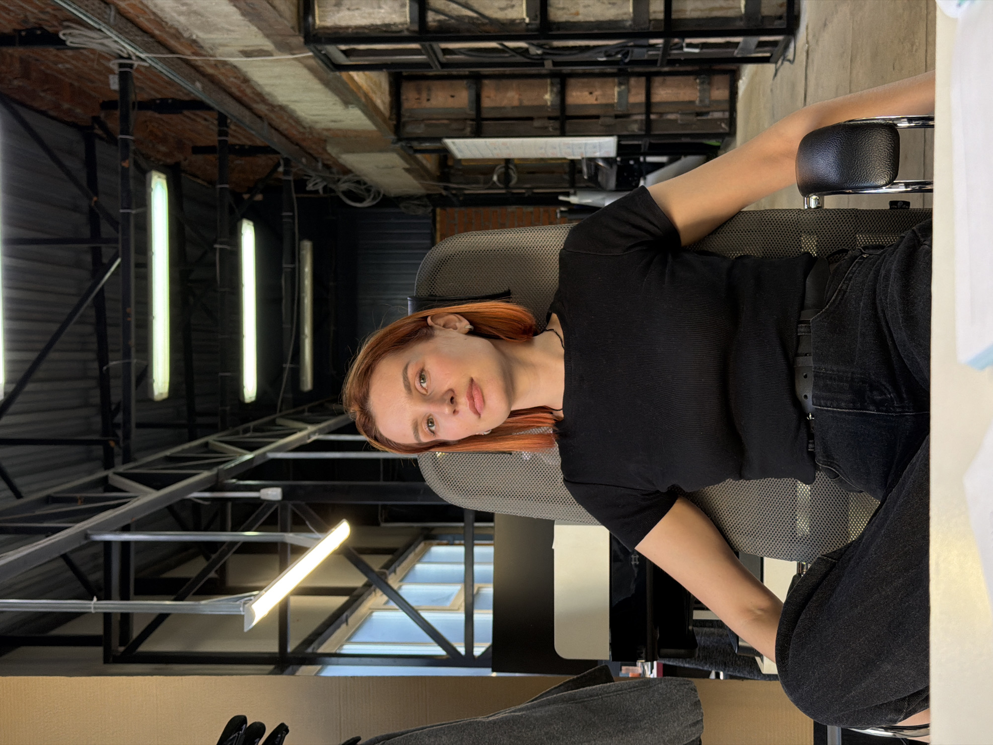 The image shows a woman with reddish-blonde hair sitting in an office chair. She is wearing a black top and dark jeans. The location appears to be an industrial-style building with exposed beams, brick, and ductwork visible in the background. There are also rows of lights illuminating the space. While the location is potentially in Chaikovsky, Russia, this is not definitively confirmed by the image's content.