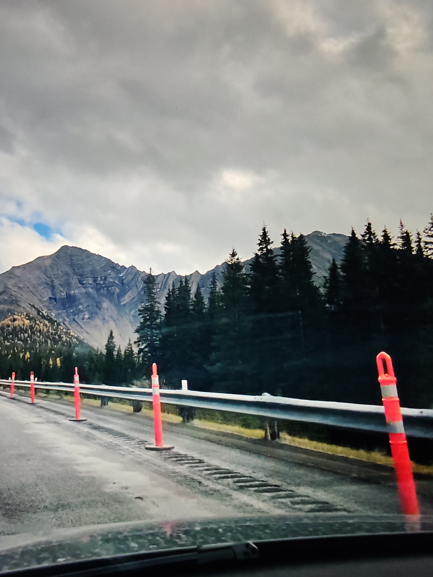 The image depicts a mountainous landscape with a road in the foreground. The road is wet and has orange traffic cones lining the right side. A metal guardrail separates the road from a grassy area with some yellow foliage. Beyond the guardrail, a dense forest of evergreen trees is visible, with a large, rugged mountain range in the background. The sky is overcast with gray clouds, though a patch of blue is visible to the left. The scene appears to be captured from a vehicle moving along the road. The location is not within the city of Calgary, Canada, but rather in a mountainous region outside of it.