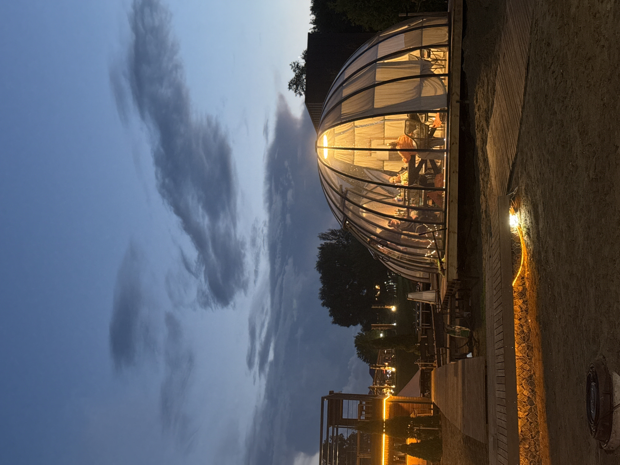 The image captures a twilight scene at a waterfront establishment in Votkinsk, Russia. The focal point is a large, geodesic dome structure with a transparent roof and walls, illuminated from within by warm lighting. Inside the dome, several individuals are seated around a table, engaged in conversation or dining. The dome is situated on a wooden platform, which extends to a wooden walkway. Adjacent to the dome is a multi-story wooden building with illuminated windows, suggesting it is part of the same establishment. The surrounding environment appears to be a sandy area, with trees and foliage visible in the background. The sky is a deep blue with scattered clouds, indicating the transition from day to night. The lighting throughout the scene, both from the interior of the dome and exterior lamps, creates a cozy and inviting atmosphere.