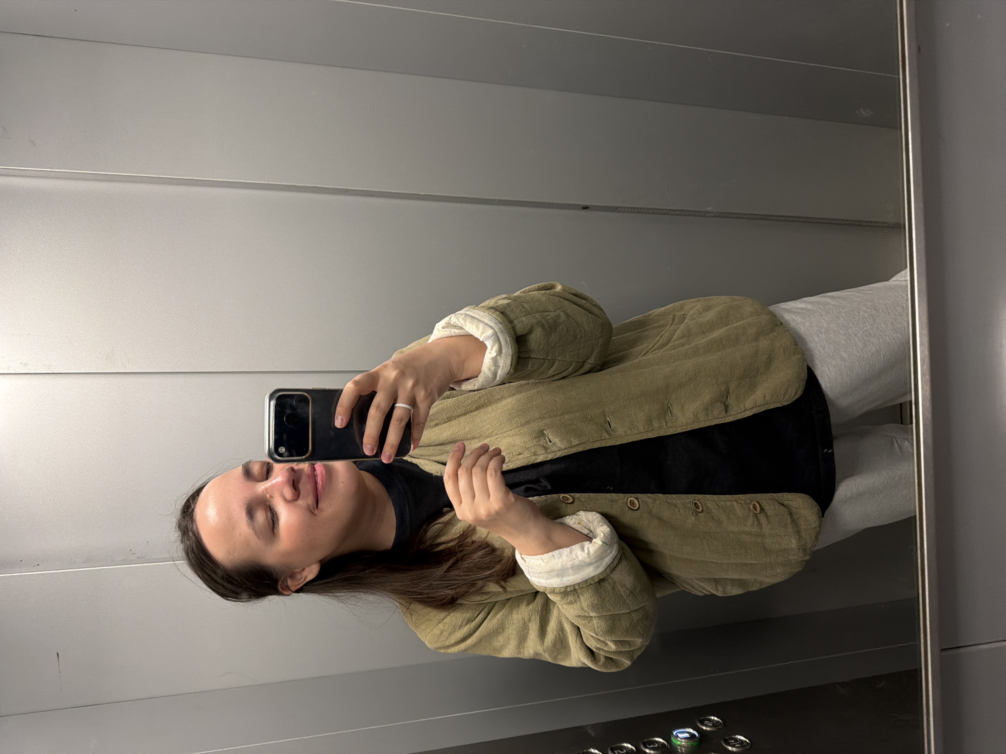 The image captures a mirror selfie taken by a person inside an elevator. The subject, a woman with long, dark brown hair, is shown from the waist up. She is wearing a casual olive-green quilted jacket with visible seams and buttons, and its cuffs are rolled up to reveal a light-colored lining. Underneath, she wears a black long-sleeved top and light grey sweatpants.
Her left hand, adorned with a simple silver-colored ring on the ring finger, holds a black smartphone with multiple camera lenses, pointed towards the mirror to capture the reflection. Her eyes are closed, and a subtle smile or serene expression is visible on her face, suggesting a relaxed moment.
The setting is an enclosed elevator space, with light grey, horizontally paneled walls that appear to be metal or a similar modern material. The right edge of the mirror's frame is visible, extending vertically. At the bottom right of the image, a panel with several metallic, round elevator buttons is partially visible, but no specific text or numbers can be discerned. The lighting is uniform and artificial, characteristic of an indoor environment. The scene appears to take place at an indeterminate time of day, within Chaikovsky, Russia.