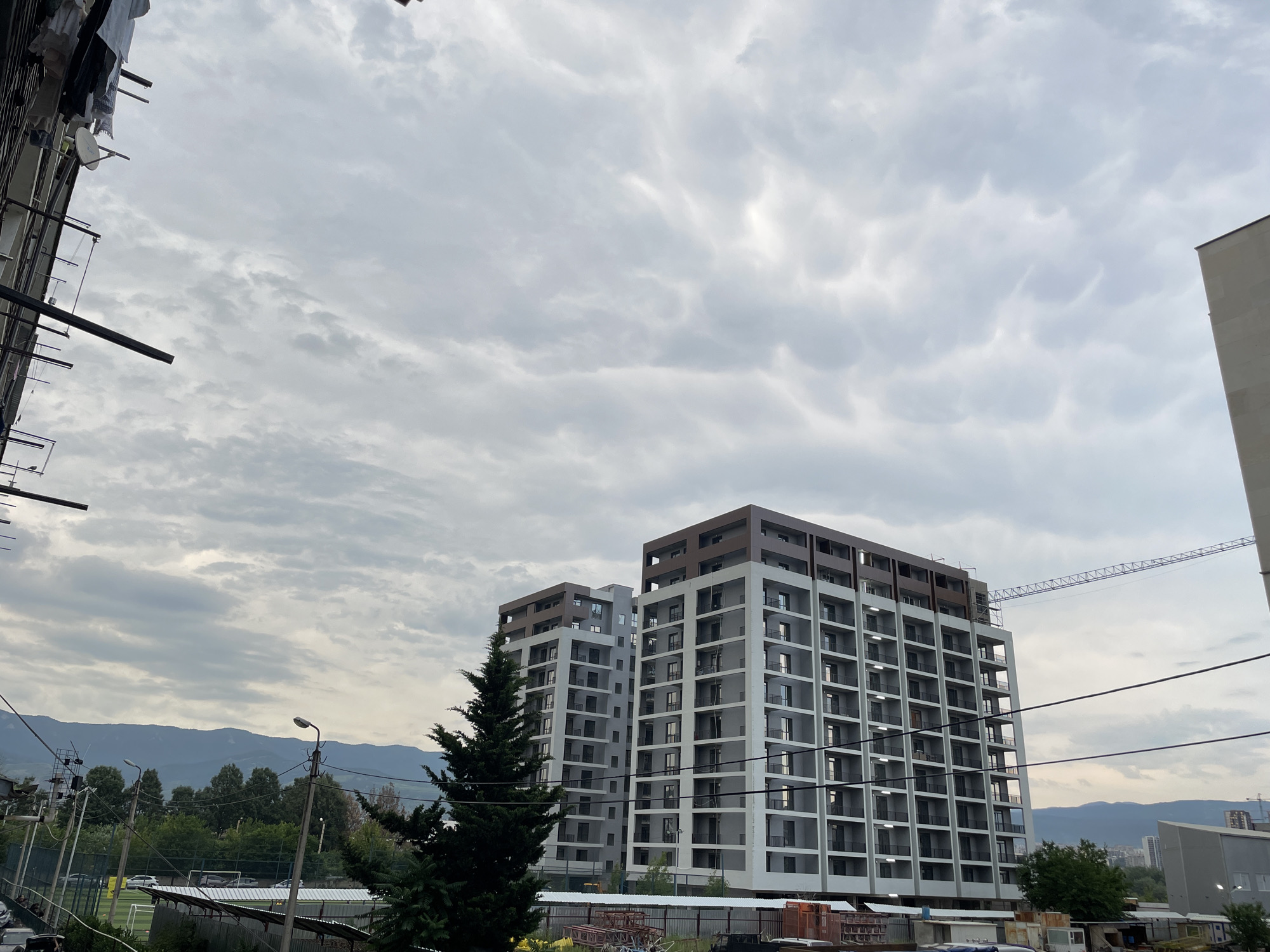 The image depicts a view of modern residential buildings under construction in Tbilisi, Georgia. In the foreground, on the left, a section of an older building is visible, with laundry hanging on lines. A tall, dark green conifer tree stands prominently in the middle ground. Behind the tree, two identical, multi-story residential buildings are situated. These buildings are characterized by their grey and white facades, with numerous square windows and balconies. The top floors of these buildings feature a darker, brown architectural detail. To the right, a construction crane is partially visible, extending from behind one of the buildings, indicating ongoing development. Further in the background, a line of trees and hills can be seen, with mountains forming a distant backdrop under a cloudy sky. The sky itself is filled with textured, gray clouds, suggesting overcast weather. A soccer field with a fence is visible on the left side of the image, suggesting the buildings are located in a developing urban area with recreational facilities. Power lines are strung across the scene, and streetlights are also present. The overall impression is one of urban growth and modernization.