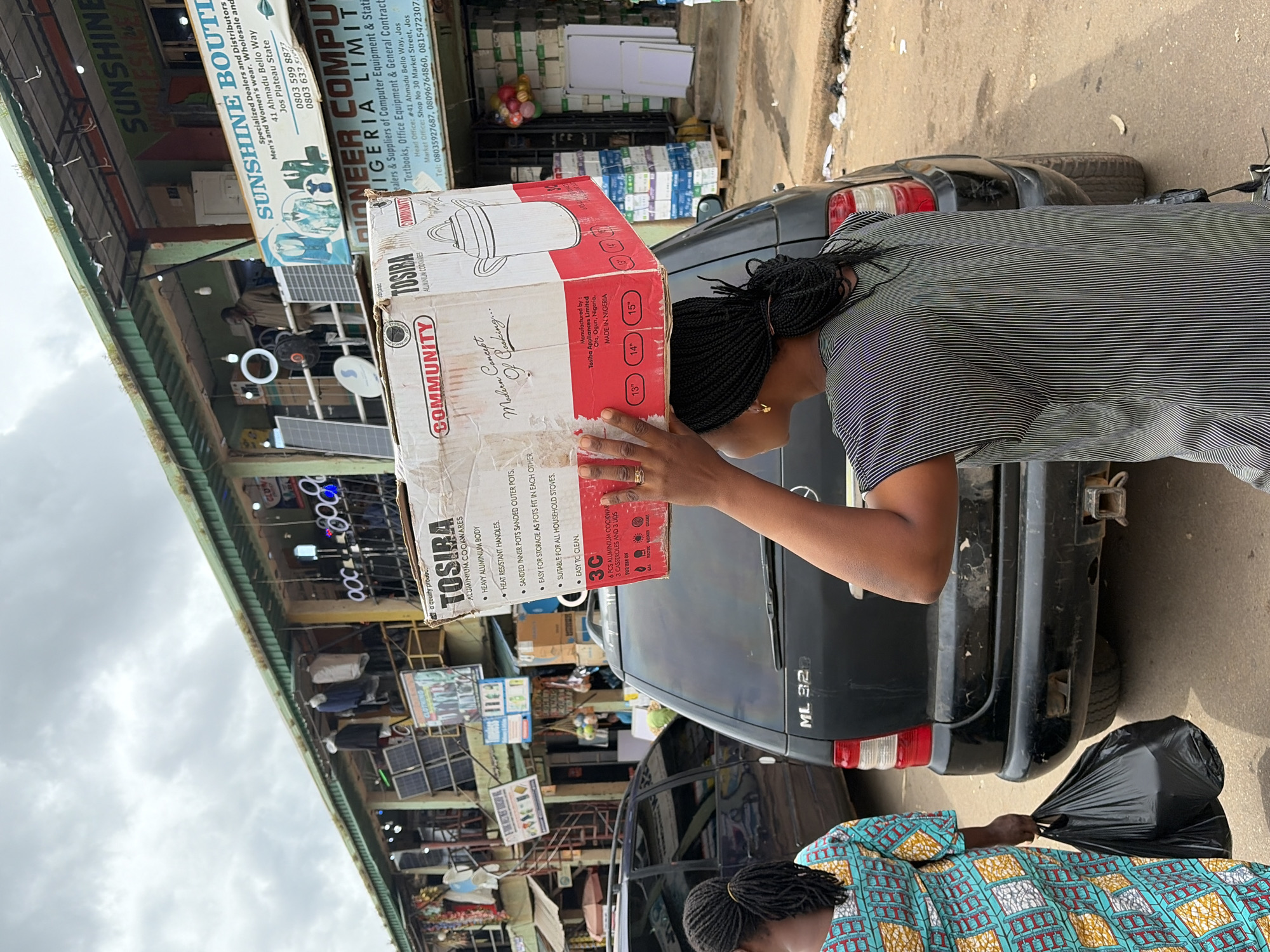 In Jos, Nigeria, a woman walks on a paved street carrying a large cardboard box of "Tosiba Aluminium Cookwares" on her head. She is wearing a striped shirt and has braided hair. Beside her, another woman in a colorful patterned dress walks with a black plastic bag in her hand. A black Mercedes-Benz ML 320 SUV is parked behind them. The background reveals a marketplace with various shops, including "Sunshine Boutique" and "Pioneer Computers Nigeria Limited". Solar panels and electrical equipment are visible on the shop fronts. The sky is overcast.