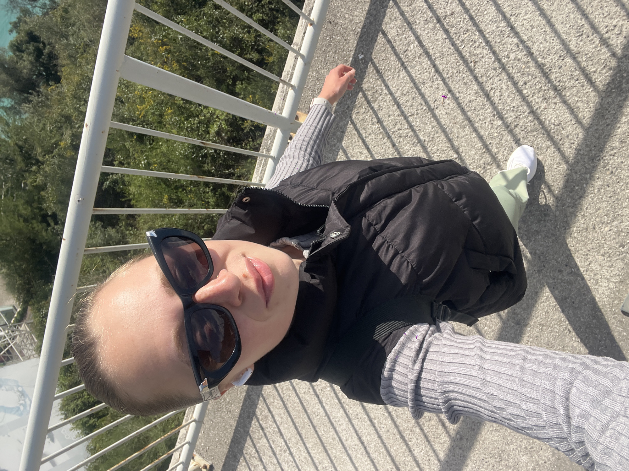 A woman stands on the Mogren 2 footbridge in Budva, Montenegro, taking a selfie. She wears sunglasses, a black puffer vest, a gray ribbed long-sleeved shirt, and light-colored pants, with a white sneaker visible. The bridge has white railings and a concrete walkway. Vegetation is visible in the background. Sunlight casts shadows from the railings onto the walkway.