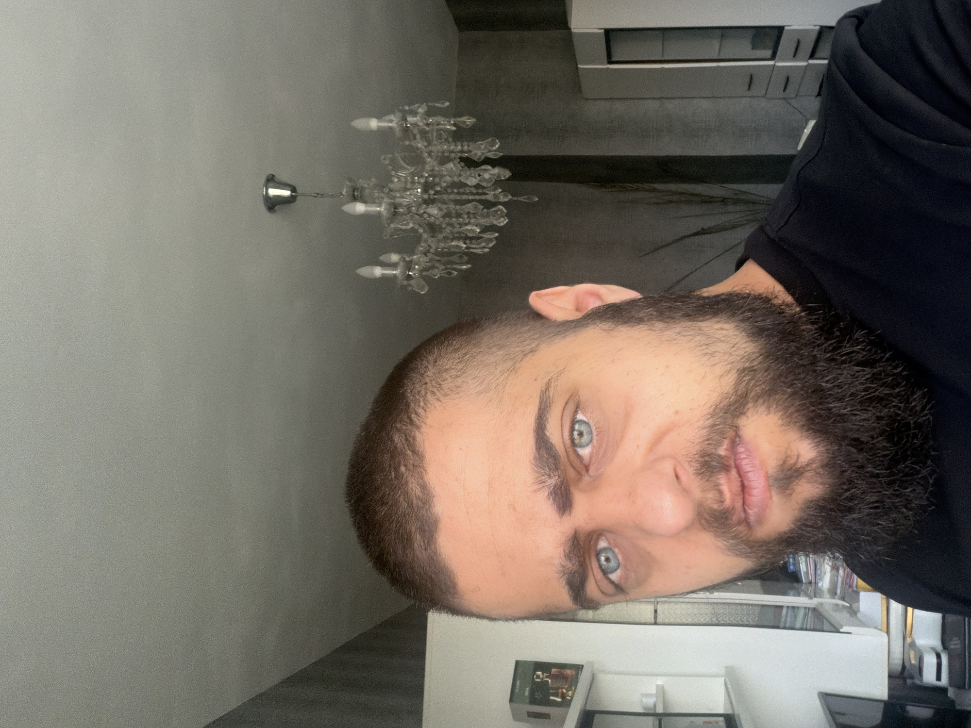A medium shot, taken from chest level, shows a man with short brown hair, a full beard, and piercing blue eyes. He is looking directly at the camera with a neutral expression. He is wearing a black t-shirt. In the background, a large crystal chandelier hangs from the ceiling. The walls are textured gray, and to the left, a white shelf unit is visible with some items on it, including a small green box. To the right, a portion of a white cabinet with black doors is visible. The lighting suggests an indoor setting, possibly a home or office.