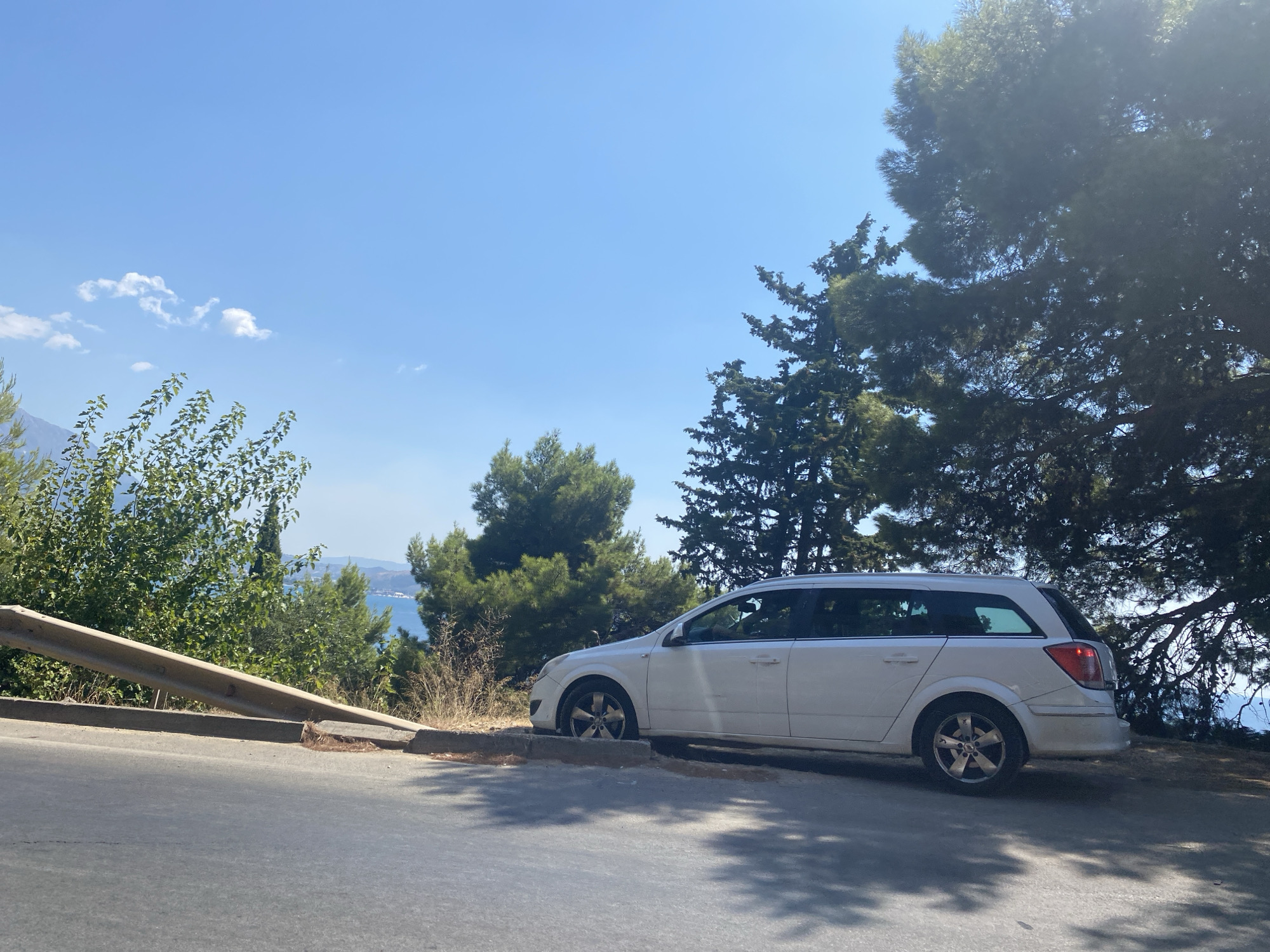 A white station wagon is parked on the side of a road overlooking a body of water, likely the Adriatic Sea, with a mountainous coastline visible in the distance. The car is positioned on a paved shoulder, with its wheels partially on a concrete curb and surrounded by dry grass and small shrubs. A metal guardrail runs along the road to the left of the car. Lush green trees, including pine trees, dominate the background, casting shadows on the road surface. The sky is a clear, bright blue with a few scattered white clouds. The scene suggests a scenic overlook or a rest stop on a coastal highway in Sutomore, Montenegro. There are no people visible in the image.