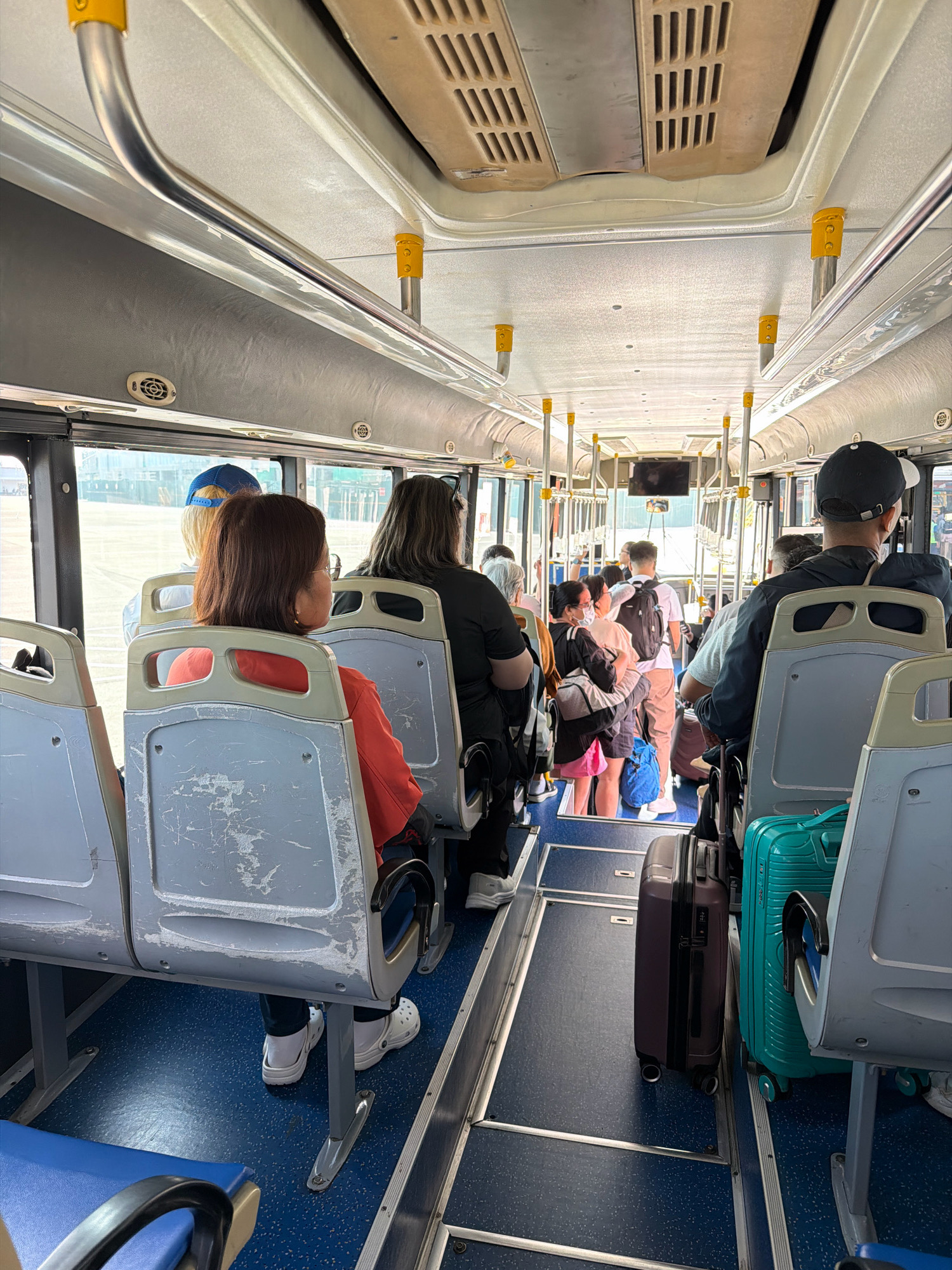 This image depicts the interior of a bus, likely a shuttle bus, carrying passengers with luggage. The bus has rows of blue and gray seats, some of which appear worn. Overhead racks and handrails are visible, along with air vents in the ceiling. Several individuals are seated and standing, with a mix of casual attire and some wearing face masks. Luggage, including suitcases and bags, is positioned throughout the aisle and near the seats. The environment suggests a functional, utilitarian mode of transportation. The presence of luggage and the bus interior points to travel, possibly to or from an airport or a tourist destination. Given the prompt's request to include the location of Nabas, Philippines, this scene could represent a shuttle service operating within the municipality, which is known for its beaches and tourist attractions such as the Malalison Island. The scene captures a moment of transit, with passengers likely on their way to or from their accommodation, the airport, or a point of interest within Nabas.