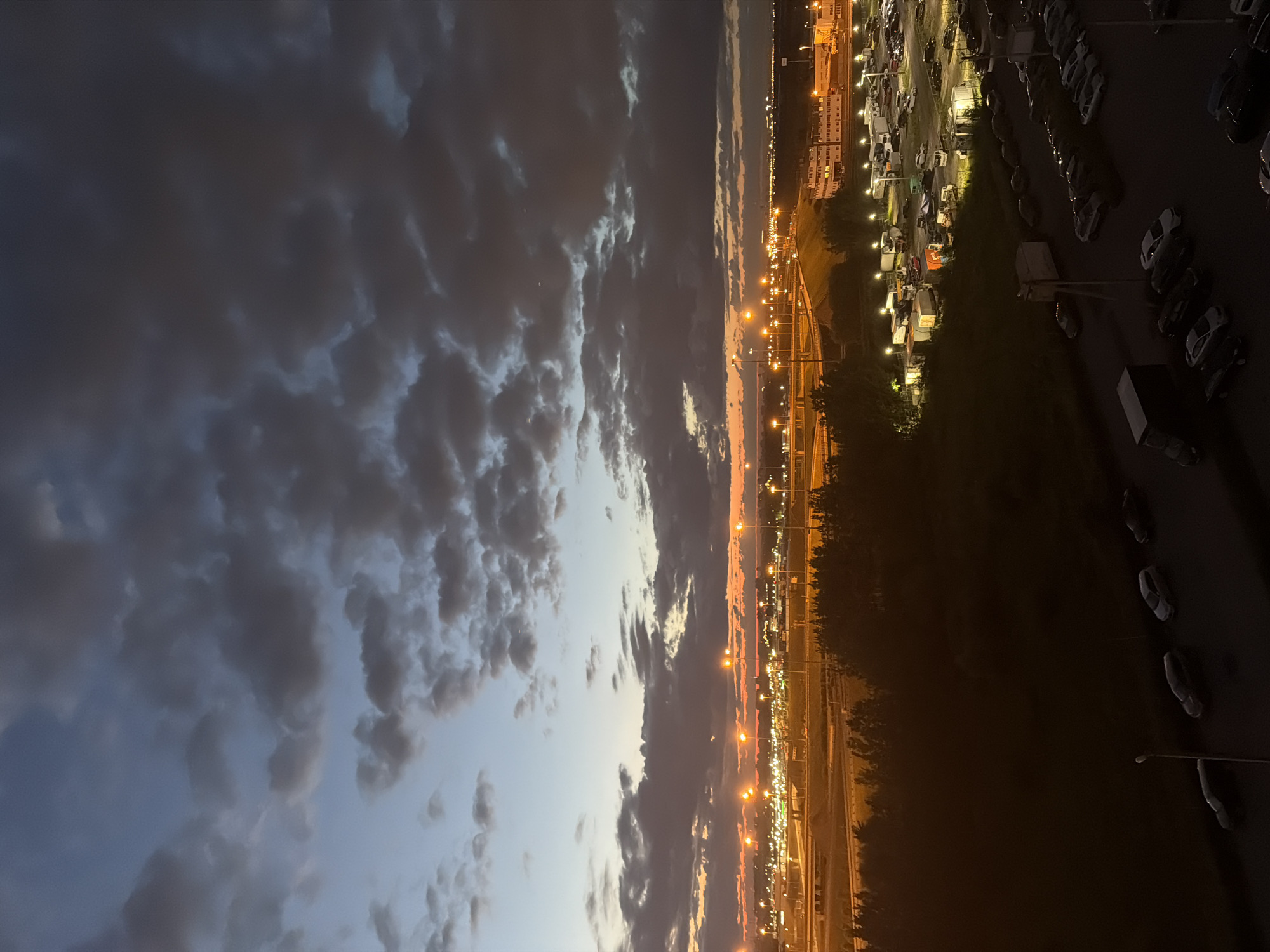 The image is a vertical, full shot of a cityscape at dusk. The sky is filled with dark, dramatic clouds that are illuminated by the fading light of the sun. The cityscape is on the right side of the image, with rows of parked cars and buildings stretching into the distance. Streetlights and lights from the buildings cast a warm glow on the scene. A road or highway runs through the middle ground, with more lights lining its path. The overall mood is peaceful and slightly melancholic, as the day transitions into night. The location appears to be a parking lot or an industrial area. There is no visible text in the image.