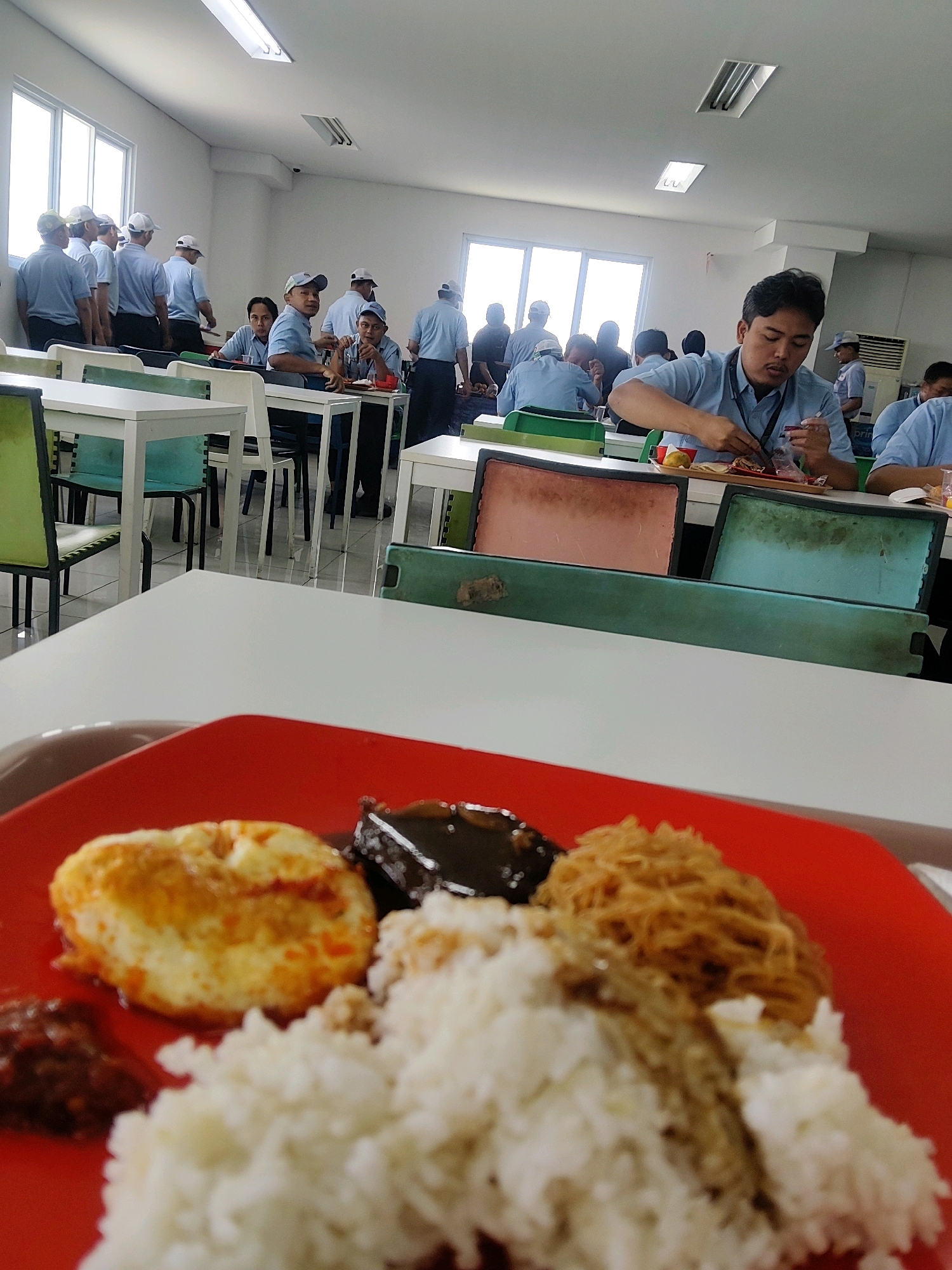 The image depicts an indoor scene, likely a cafeteria or dining hall, in Mekarmukti, Indonesia. In the foreground, a red plate holds a meal consisting of white rice, a fried egg, a dark sauce, and a portion of shredded fried material. The background shows numerous individuals, predominantly men, seated at tables and dining. Many are wearing blue shirts and white caps, suggesting they are employees or students. The environment appears to be a large, well-lit room with white walls and ceiling, and tables and chairs arranged for communal dining. Large windows are visible on one side of the room, letting in natural light. The overall impression is of a busy, functional space where people are gathered for a meal.
